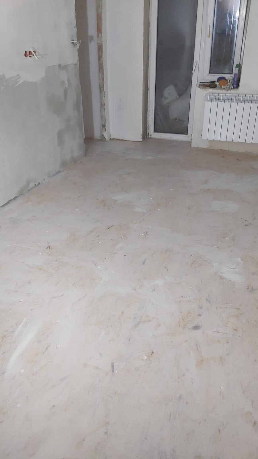 Przygotowane do dalszych prac wykończeniowych pomieszczenie z surowymi ścianami, gładkim betonowym podkładem podłogowym i grzejnikiem pod oknem.