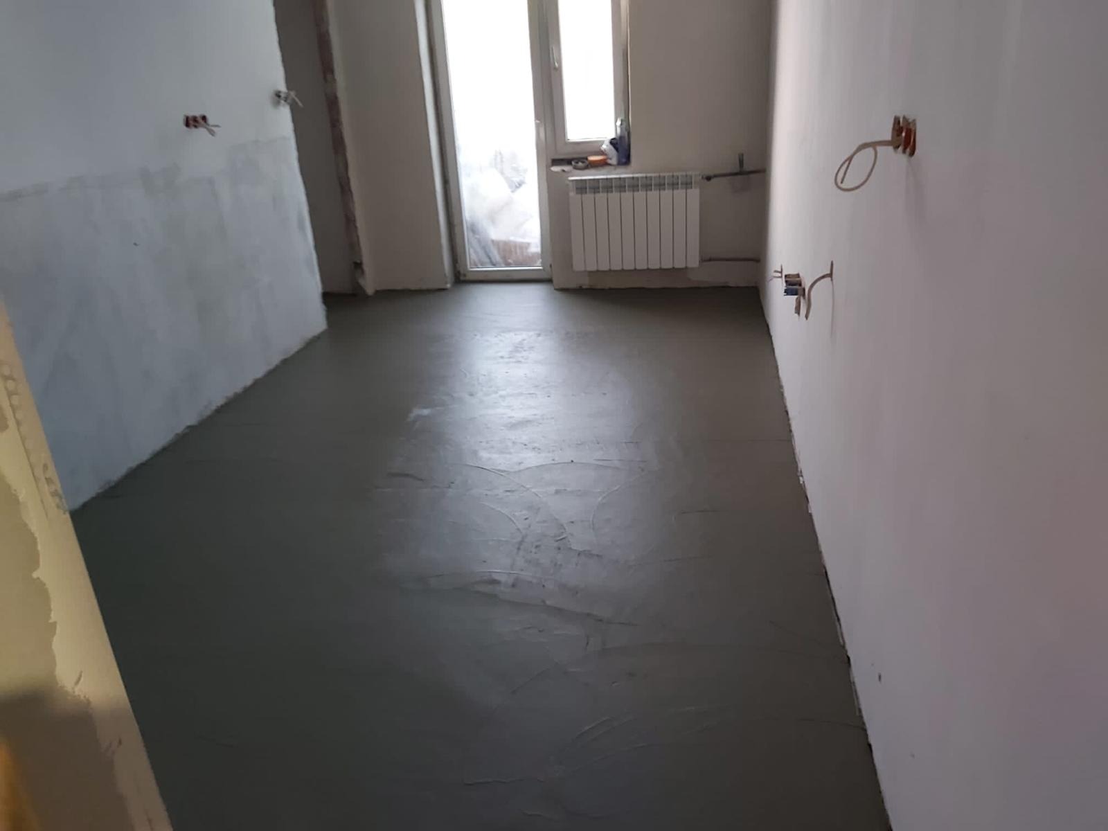 Wylany, świeży podkład podłogowy w pomieszczeniu z widocznymi puszkami elektrycznymi i grzejnikiem pod oknem.