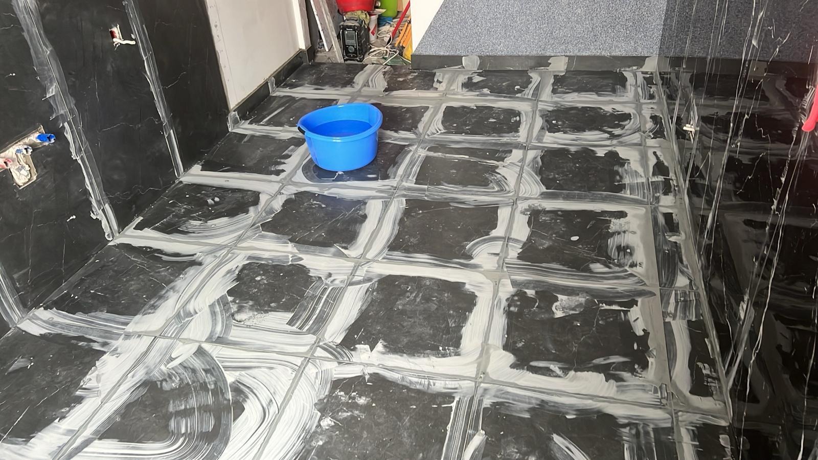 Świeżo położone, czarne płytki podłogowe z marmurkowym wzorem, w trakcie fugowania białą zaprawą, z niebieskim wiadrem z wodą w rogu pomieszczenia.