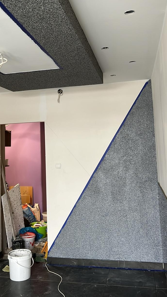 Wnętrze w trakcie prac wykończeniowych: ściana z fragmentem pokrytym szarą strukturą, zabezpieczona niebieską taśmą malarską, sufit z dekoracyjnym podwieszanym elementem również pokrytym szarą...