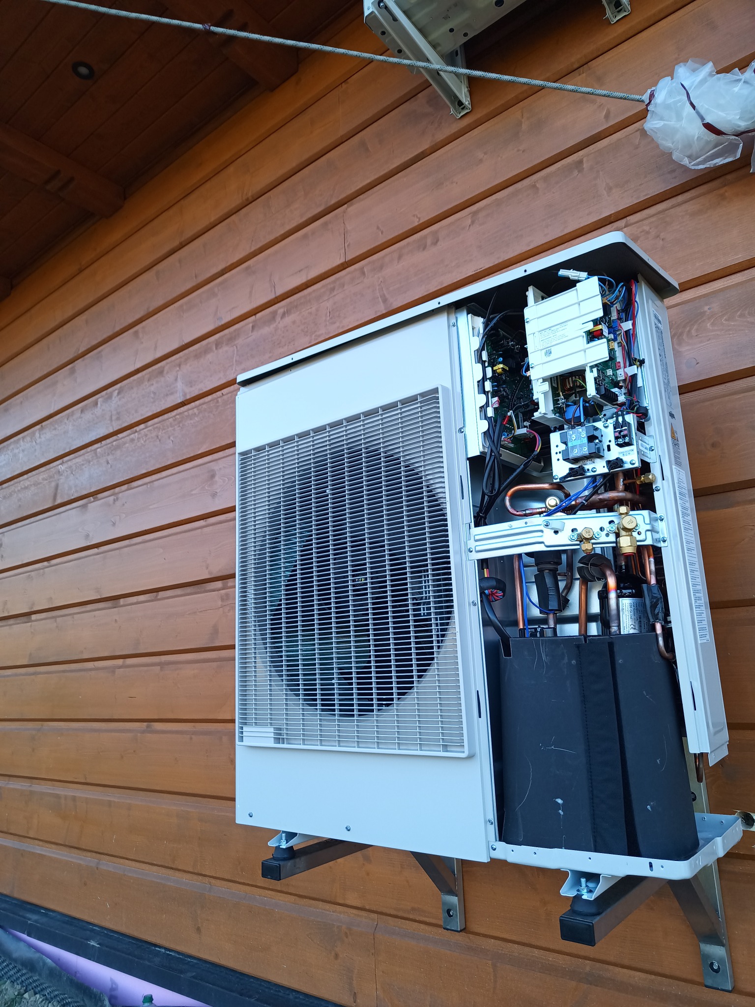 Zamontowana na drewnianej elewacji zewnętrzna jednostka pompy ciepła z otwartą obudową, widoczne wewnętrzne komponenty, rury miedziane i okablowanie.