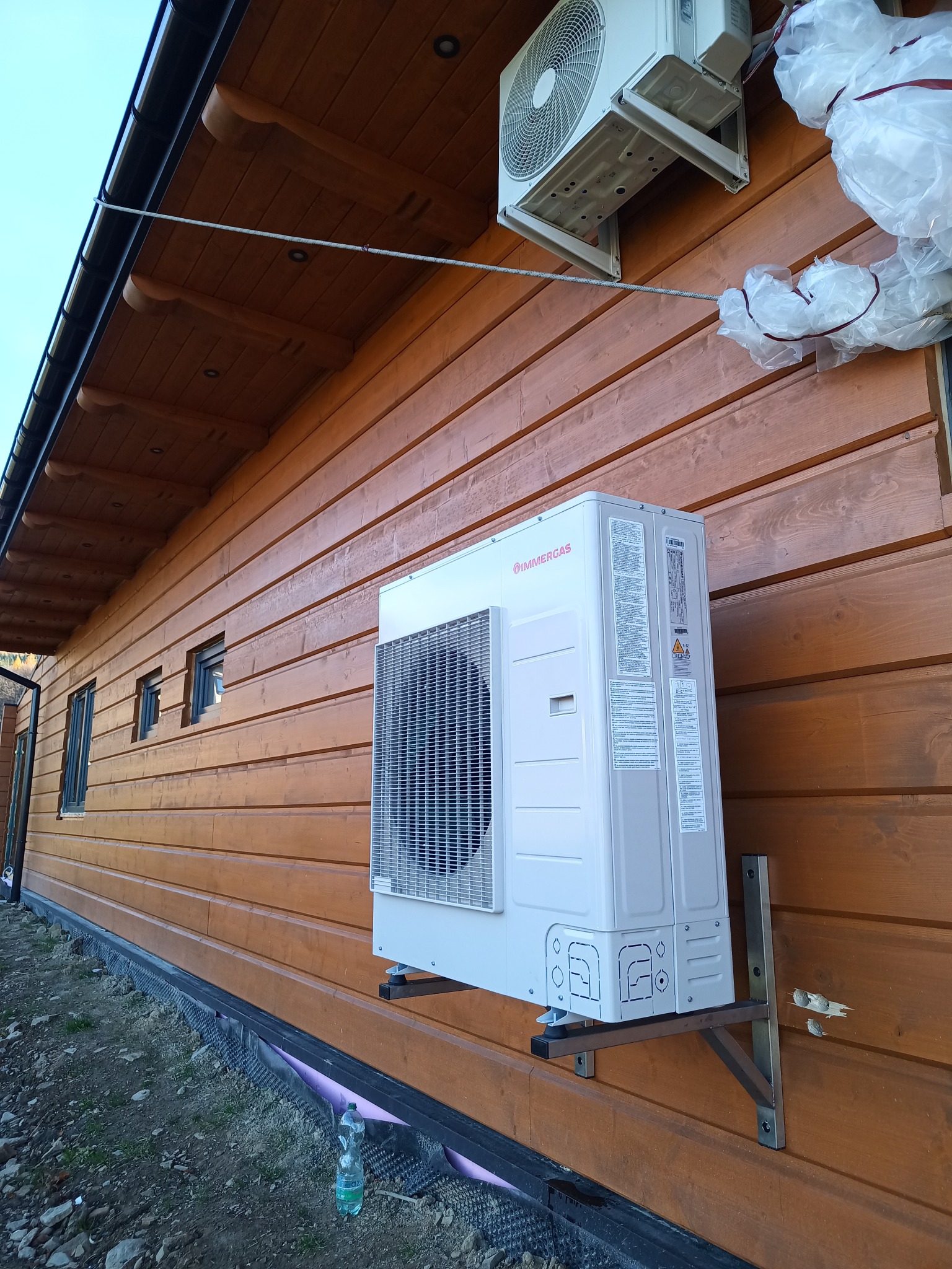 Dwa klimatyzatory marki Immergas zamontowane na drewnianej elewacji domu, jeden częściowo owinięty folią ochronną, widoczne metalowe wsporniki montażowe i butelka wody u podstawy ściany.