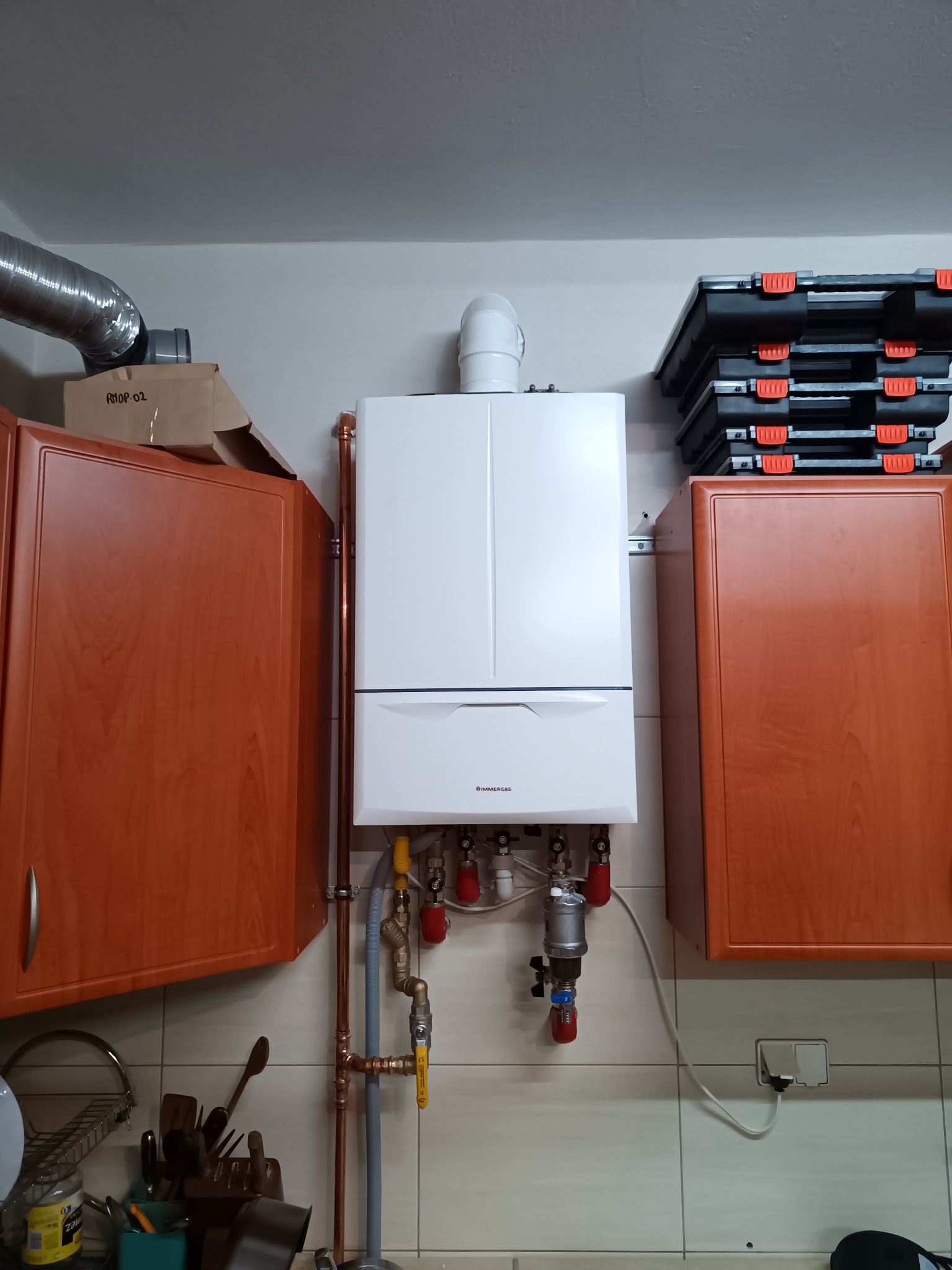 Biały piec gazowy Immergas zamontowany na ścianie z beżowych płytek, z widocznymi miedzianymi rurami, zaworami z czerwonymi pokrętłami i filtrem, otoczony brązowymi szafkami kuchennymi i stertą...