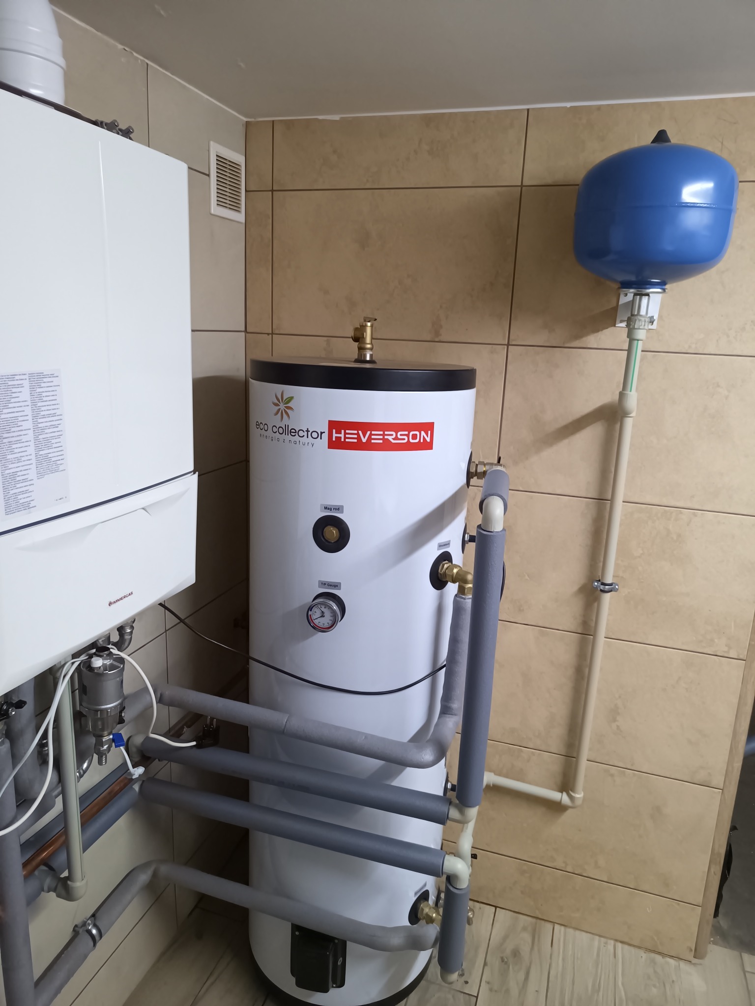 Biały piec Immergas i biały zasobnik ciepłej wody Eco Collector Heverson z szarymi rurami izolowanymi, niebieskie naczynie przeponowe, zamontowane w pomieszczeniu z płytkami imitującymi drewno.