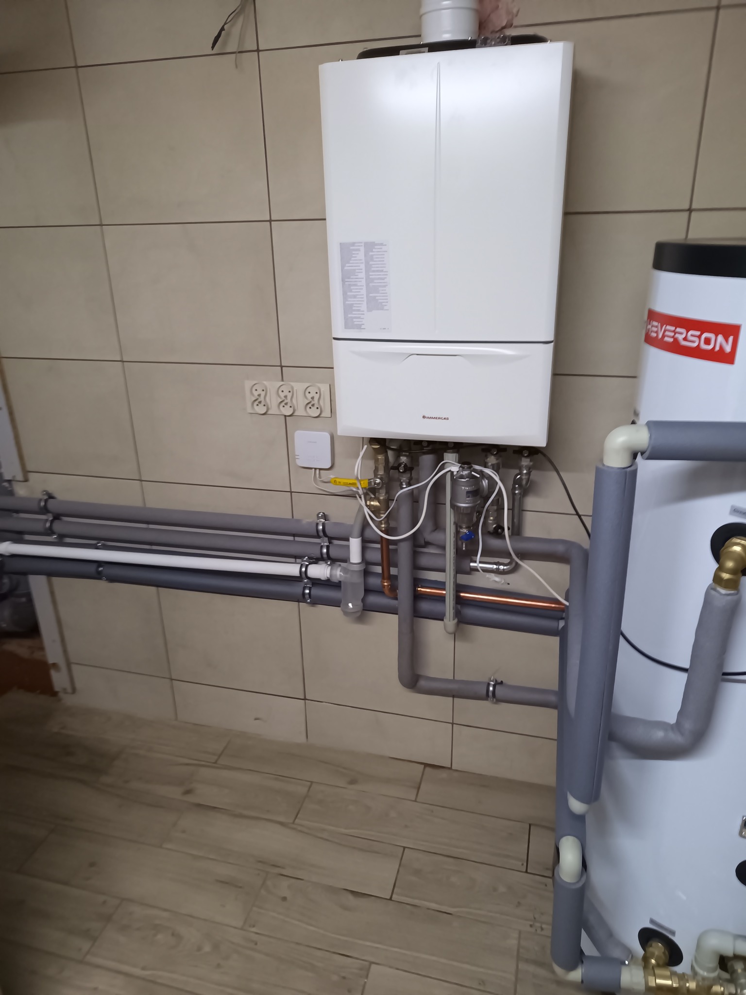 Kocioł grzewczy marki Immergas zamontowany na ścianie z beżowych płytek, widoczne rury instalacji centralnego ogrzewania, zasobnik ciepłej wody użytkowej Heverson, biały sterownik temperatury...