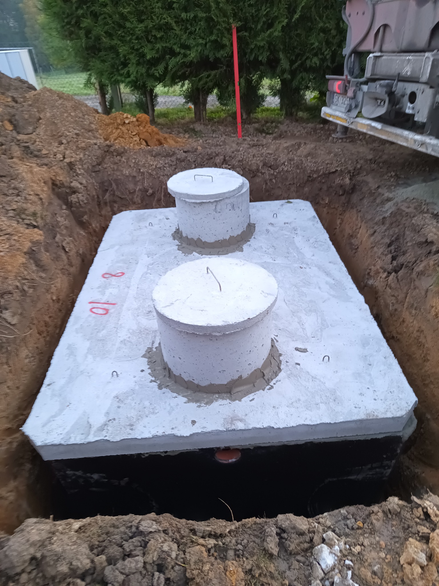 Betonowy zbiornik, prawdopodobnie szambo, umieszczony w wykopie ziemnym, z widocznymi dwoma włazami i numerami '10 8' namalowanymi czerwoną farbą na bocznej ścianie zbiornika; w tle fragment...