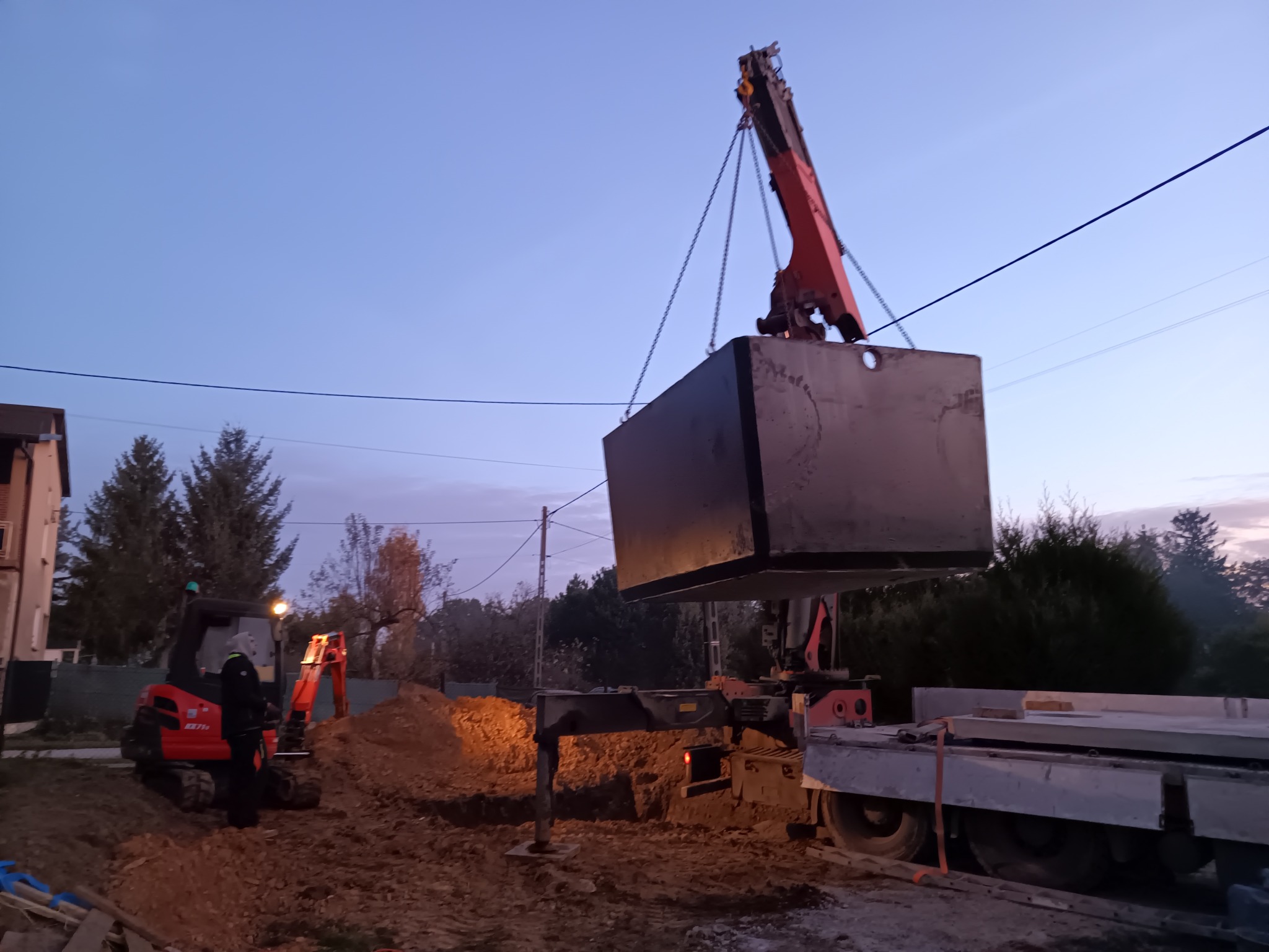 Montaż betonowego zbiornika za pomocą dźwigu na placu budowy, widoczna koparka i wykop w ziemi.