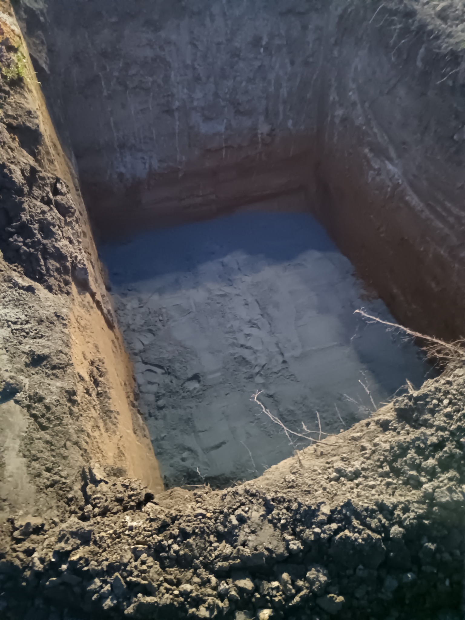 Wykop o kwadratowym kształcie z pionowymi ścianami, wykonany w ziemi o różnej strukturze i kolorystyce, oświetlony z góry, widoczne korzenie na krawędzi.