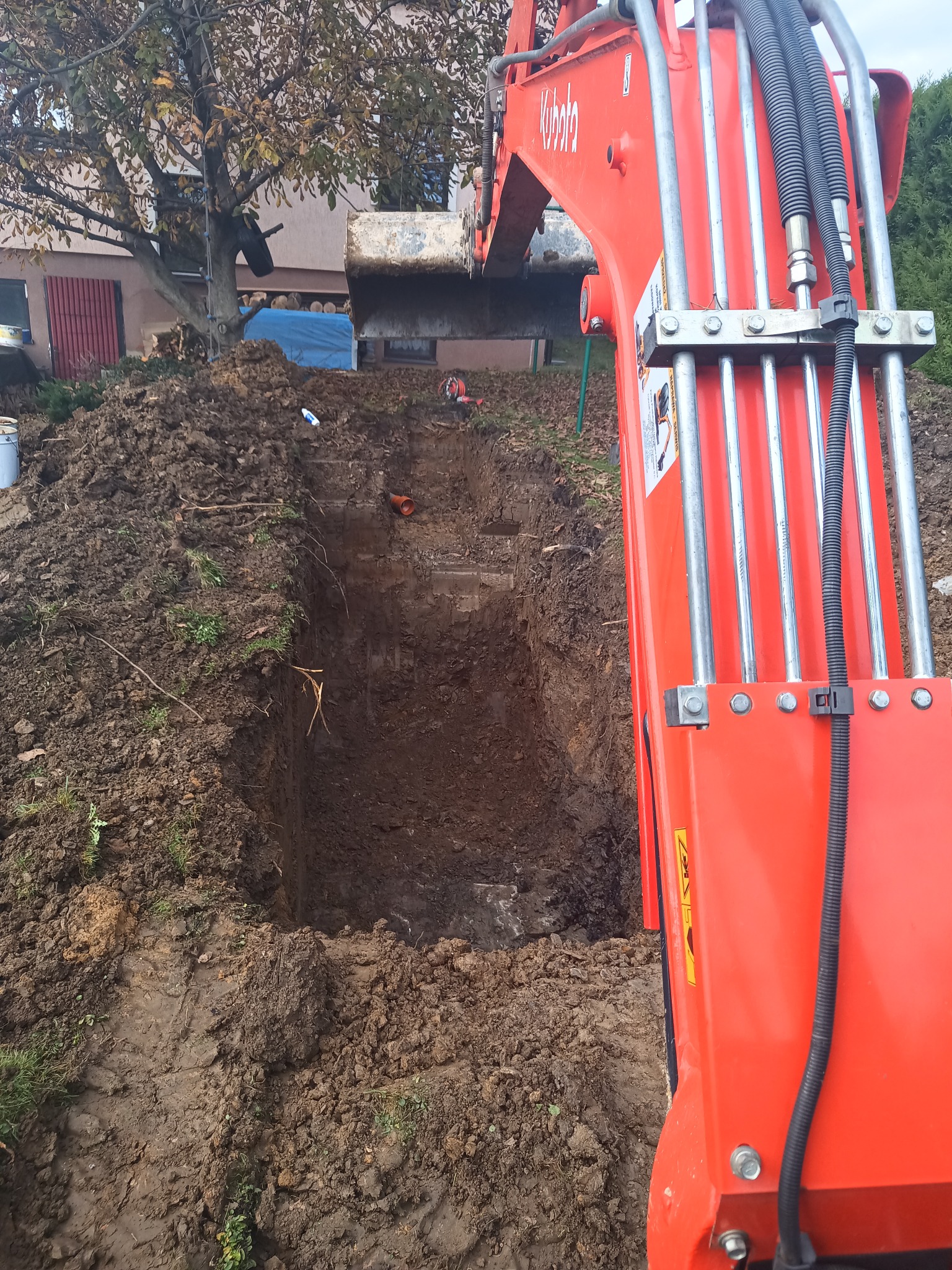 Pomarańczowa koparka Kubota w trakcie wykopywania głębokiego wykopu w ziemi, widoczne przewody hydrauliczne, w tle budynek mieszkalny i drzewa