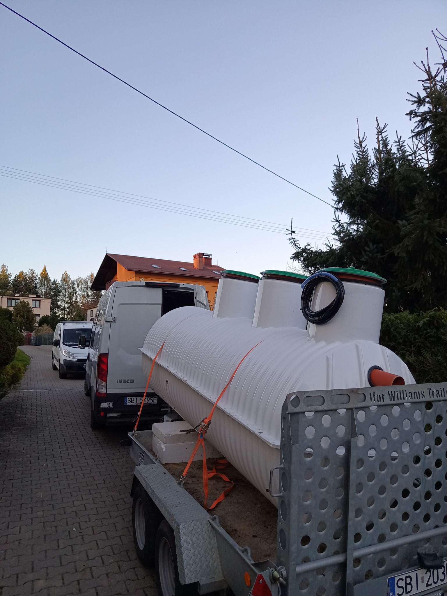 Biały zbiornik z trzema wlotami, zabezpieczony pomarańczowymi pasami, transportowany na przyczepie za Iveco, widok z boku na tle zabudowań i drzew.