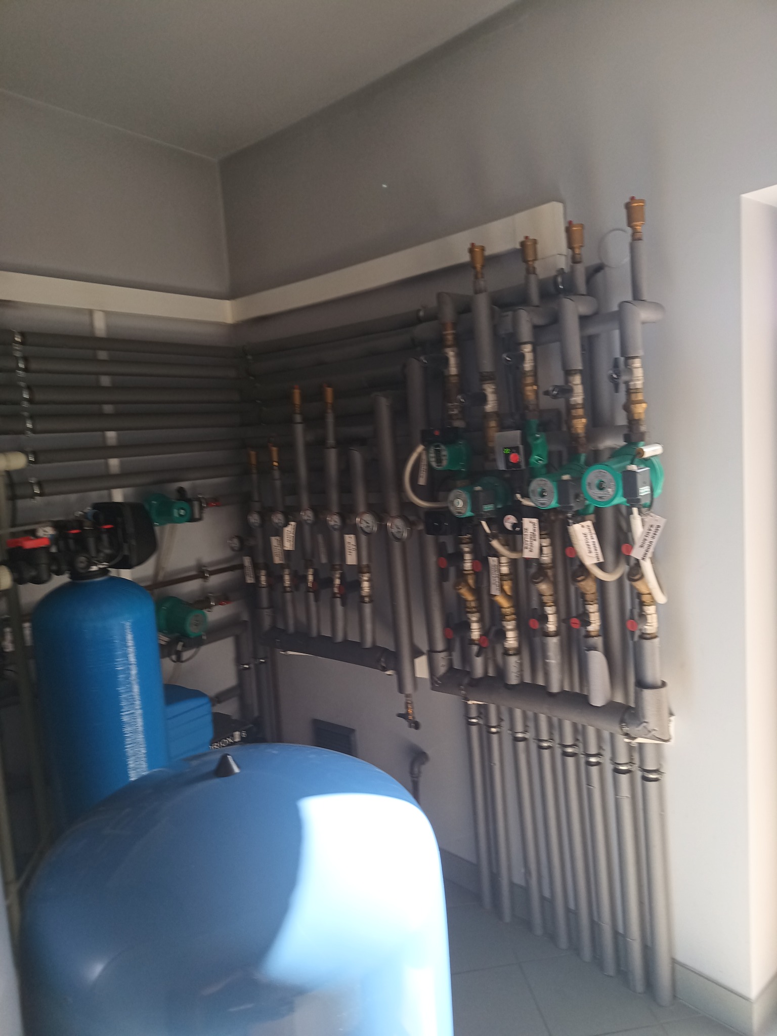 Zestaw instalacji centralnego ogrzewania z niebieskimi zbiornikami hydroforowymi, licznymi rurami w izolacji i pompami obiegowymi, widoczne liczniki ciepła i manometry.