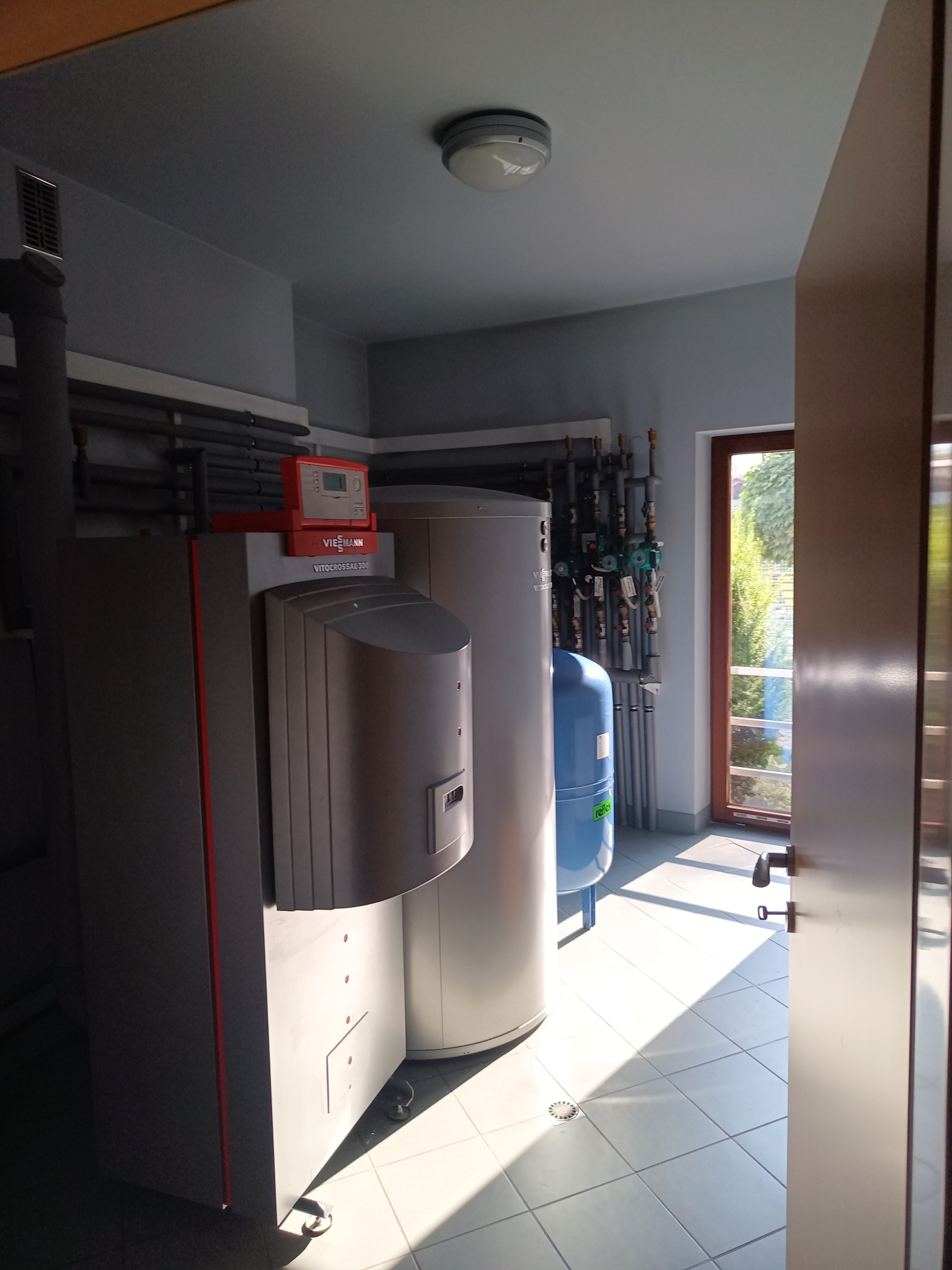 Kotłownia z kotłem gazowym Viessmann Vitocrossal 300, zbiornikiem akumulacyjnym, niebieskim naczyniem wzbiorczym Reflex i instalacją rurową, widoczne przez otwarte drzwi przy jasnej podłodze z płytek.