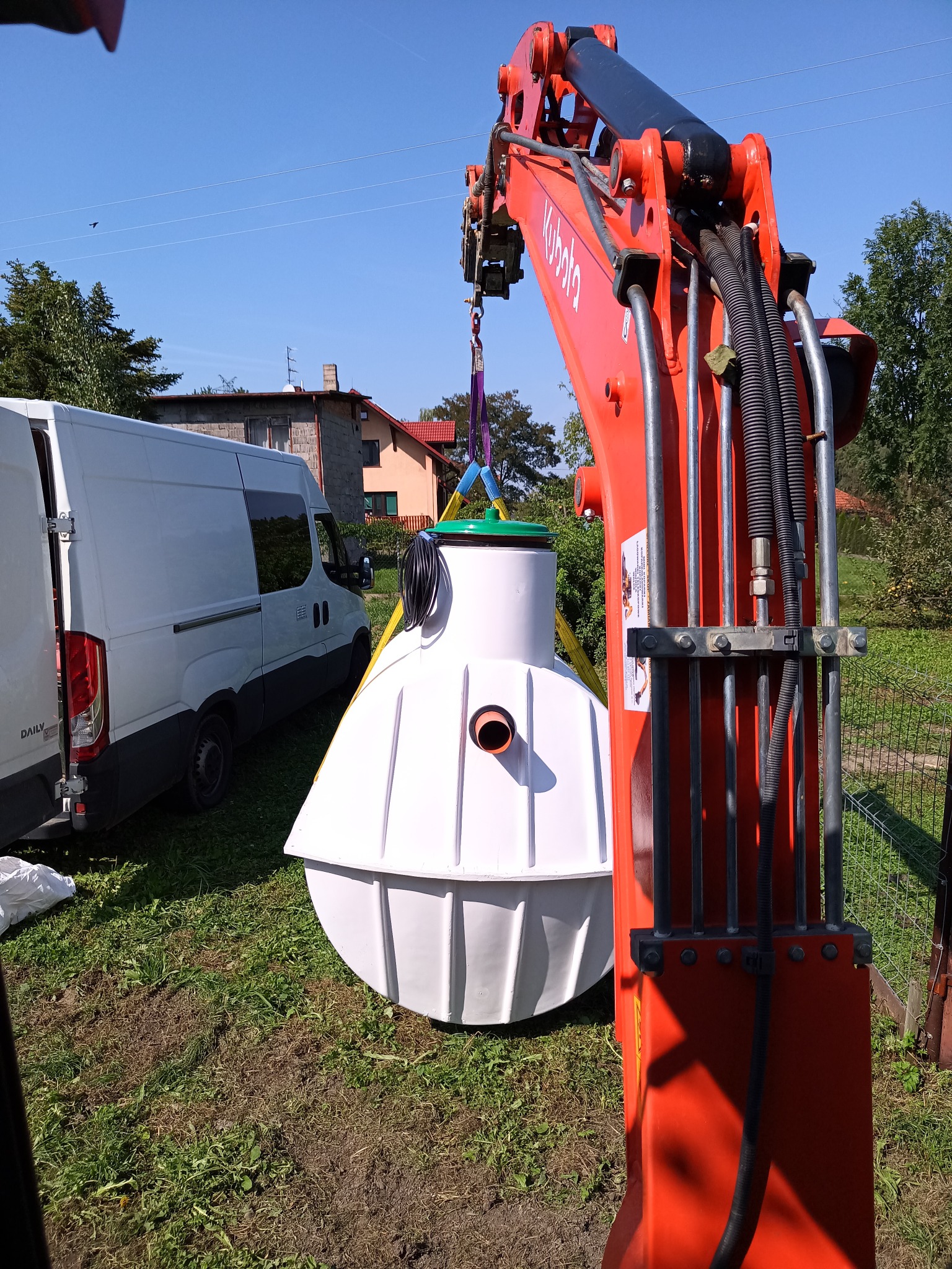 Montaż przydomowej oczyszczalni ścieków: biały zbiornik z zieloną pokrywą zawieszony na haku koparki Kubota, w tle biały van i budynek mieszkalny.