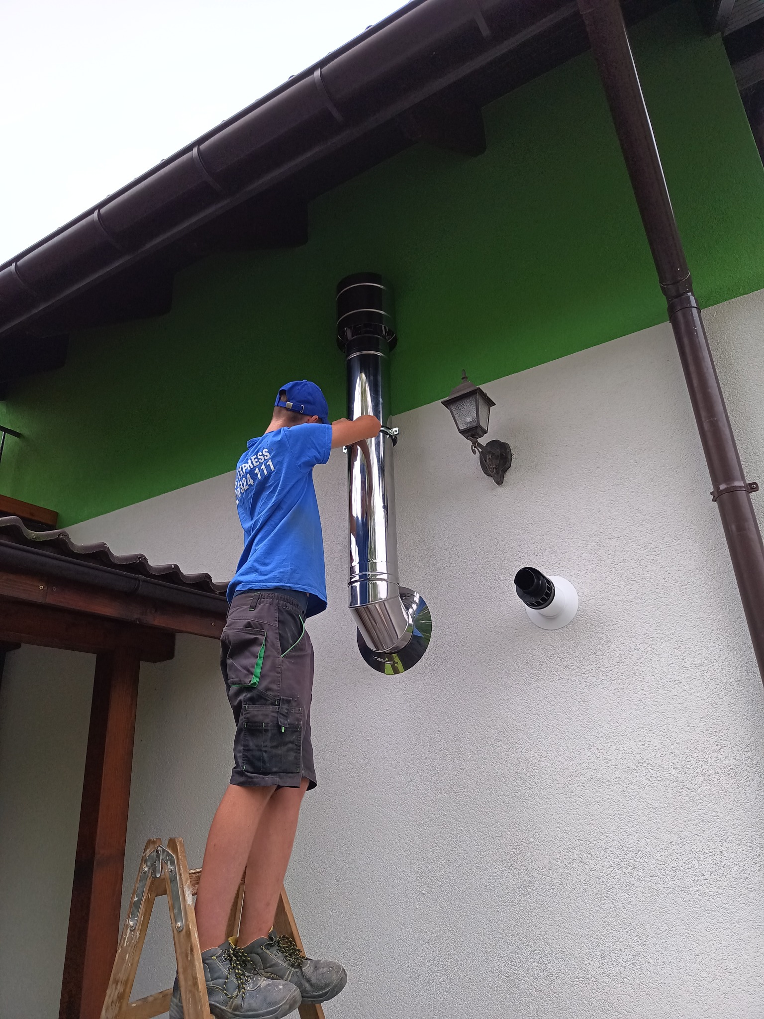 Monter na drabinie instaluje błyszczący, metalowy komin na zewnątrz budynku z zielonym pasem pod brązowym dachem, obok lampy i kamery.