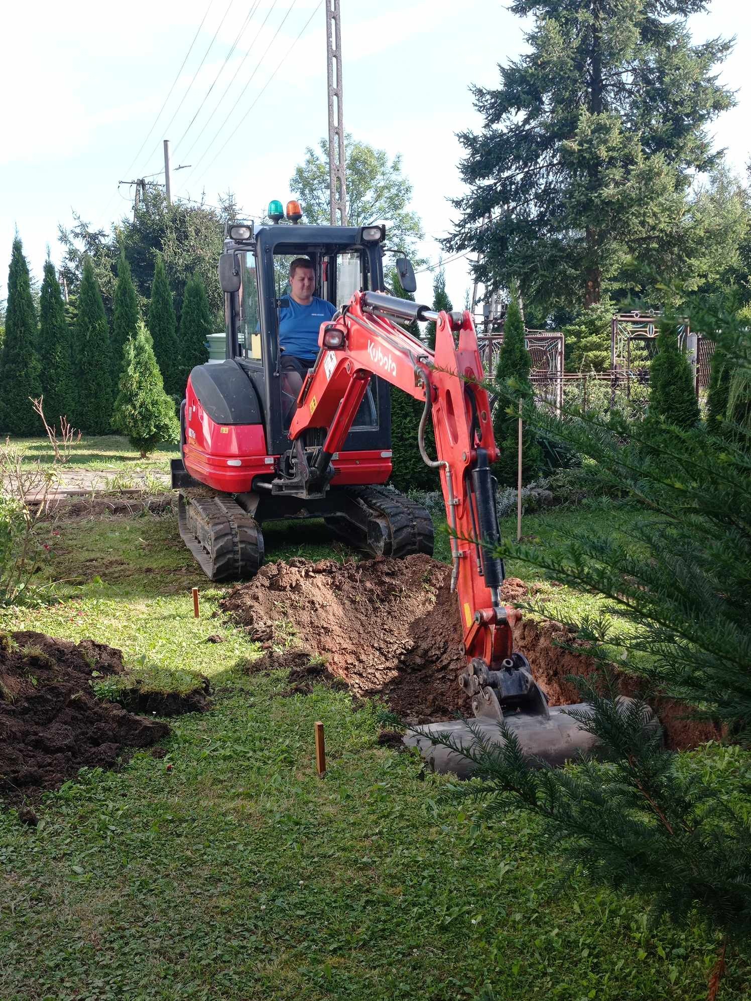 Operator w kabinie czerwonej mini-koparki Kubota podczas prac ziemnych na trawniku, widoczne wykopane zagłębienie i paliki wyznaczające trasę.