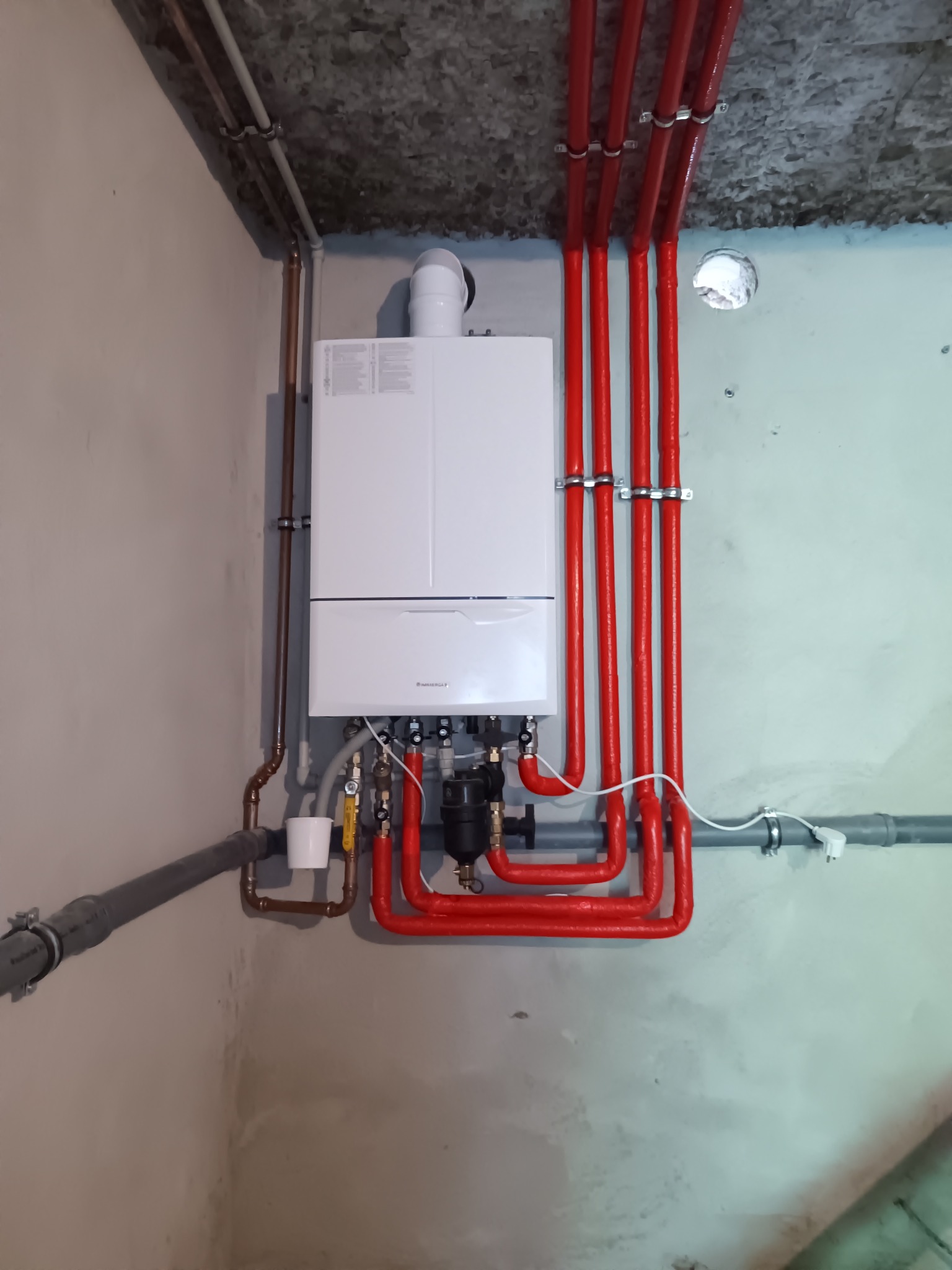 Nowoczesny, biały piec centralnego ogrzewania zamontowany na szarej ścianie, z widocznymi czerwonymi rurami instalacji grzewczej i miedzianymi rurami przyłączeniowymi, w surowym pomieszczeniu...