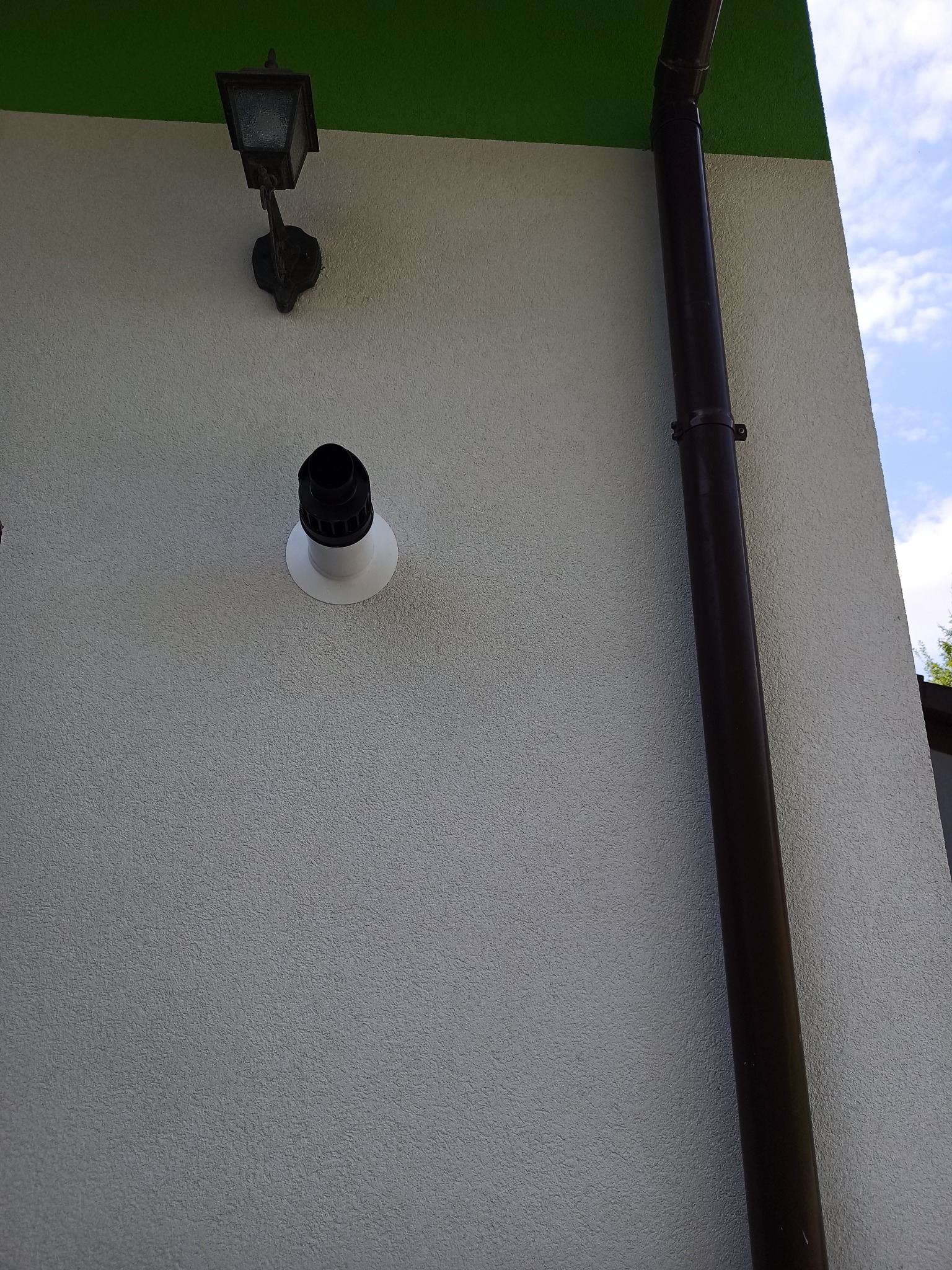 Fragment ściany budynku z zamontowanym odpowietrznikiem kanalizacyjnym, rynną spustową i lampą zewnętrzną.