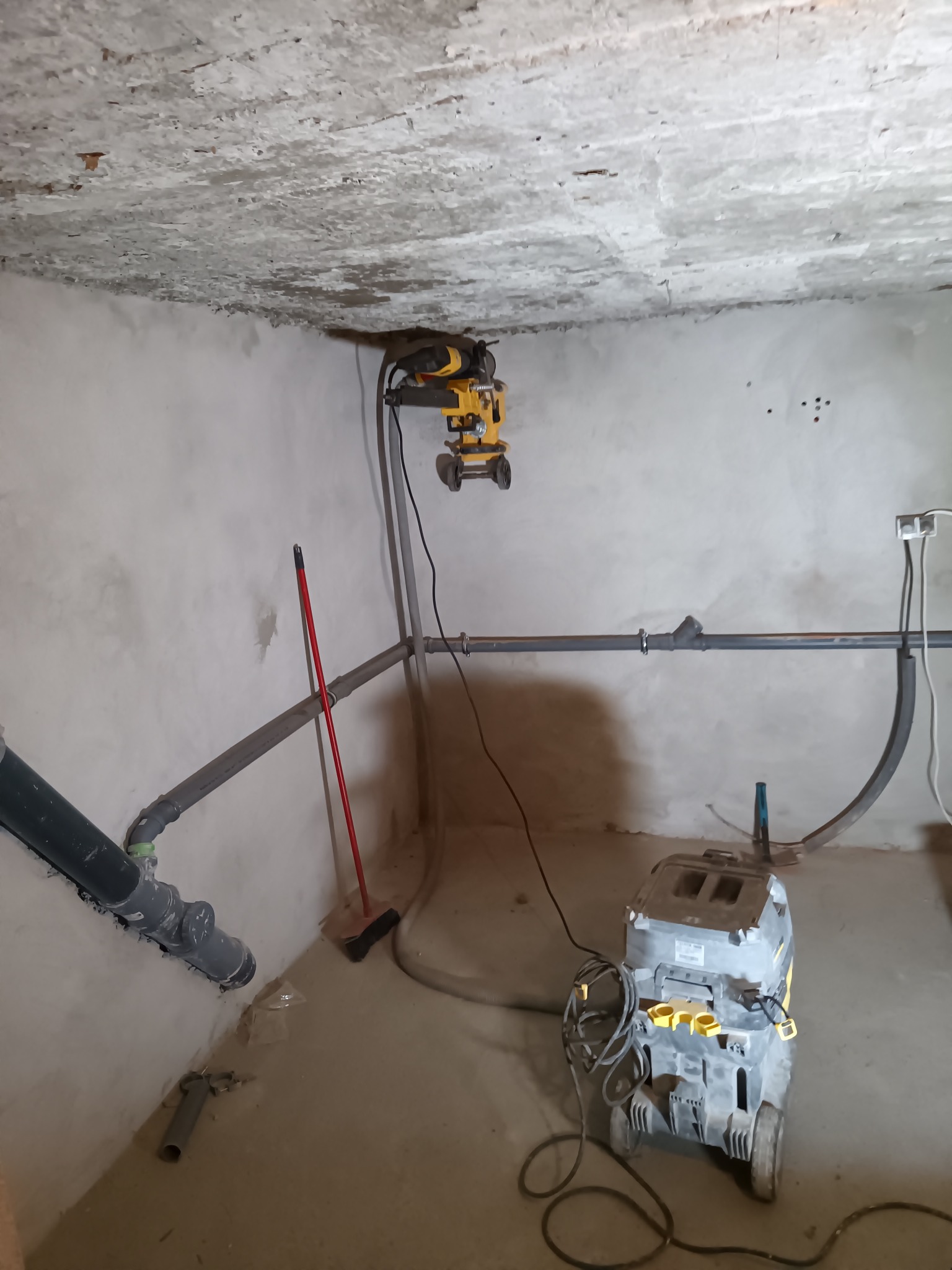 Szare rury kanalizacyjne zamontowane w surowym pomieszczeniu z betonowym sufitem, widoczny odkurzacz przemysłowy i narzędzie do wiercenia w betonie.