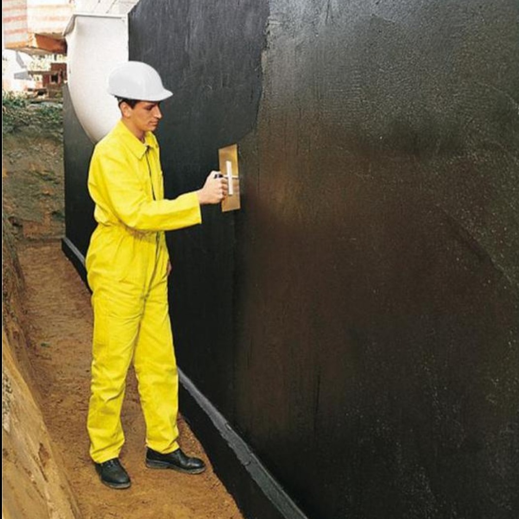 Pracownik w żółtym kombinezonie i białym kasku aplikuje czarną, gęstą substancję na ścianę fundamentu za pomocą pacy. Widoczne ślady świeżo nałożonej warstwy.