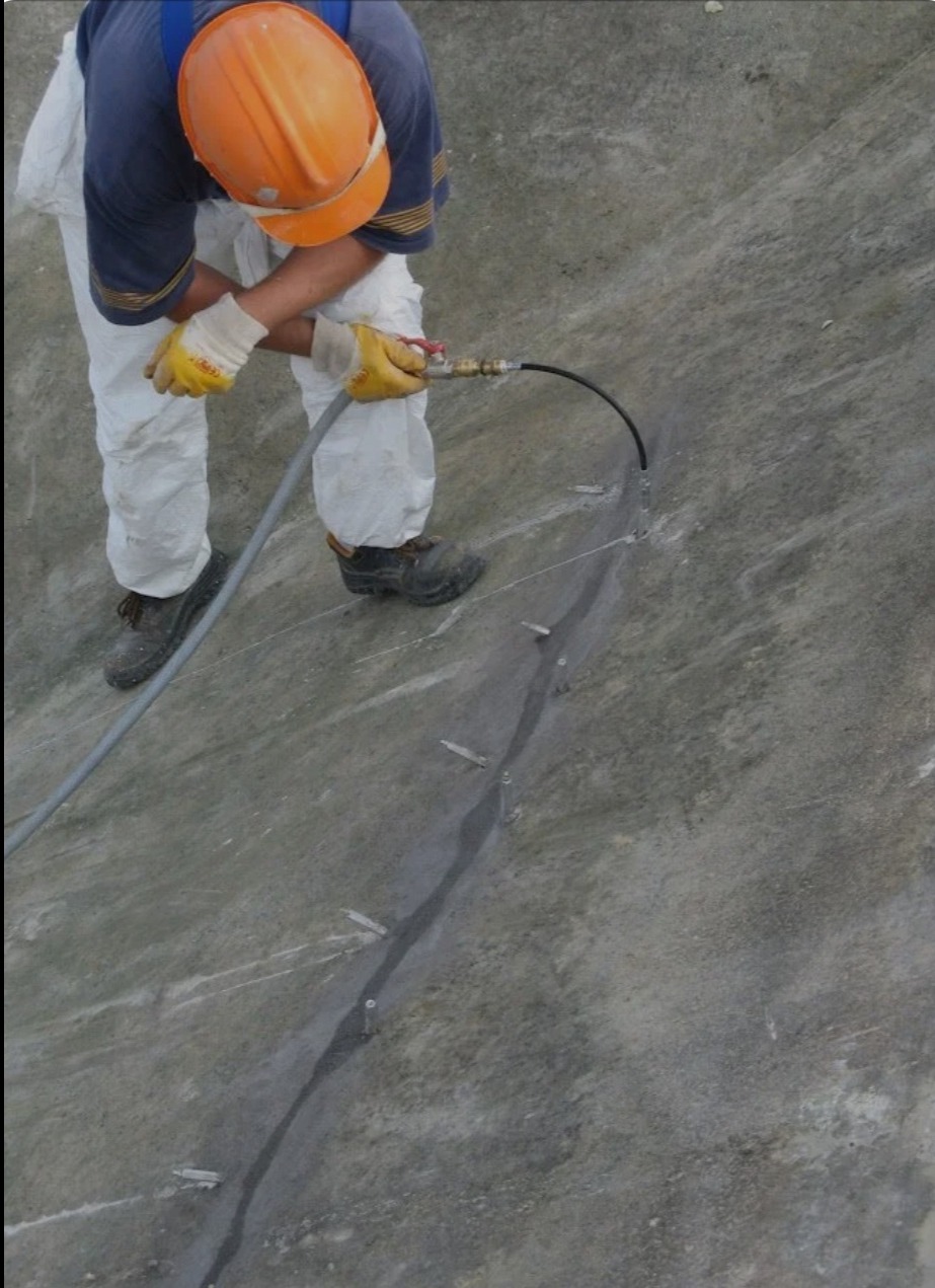 Pracownik w kombinezonie i kasku aplikuje uszczelniacz do szczeliny w betonowej powierzchni, prawdopodobnie fundamentu, używając węża z dyszą. Widoczne ślady wcześniejszych napraw i elementy mocujące.