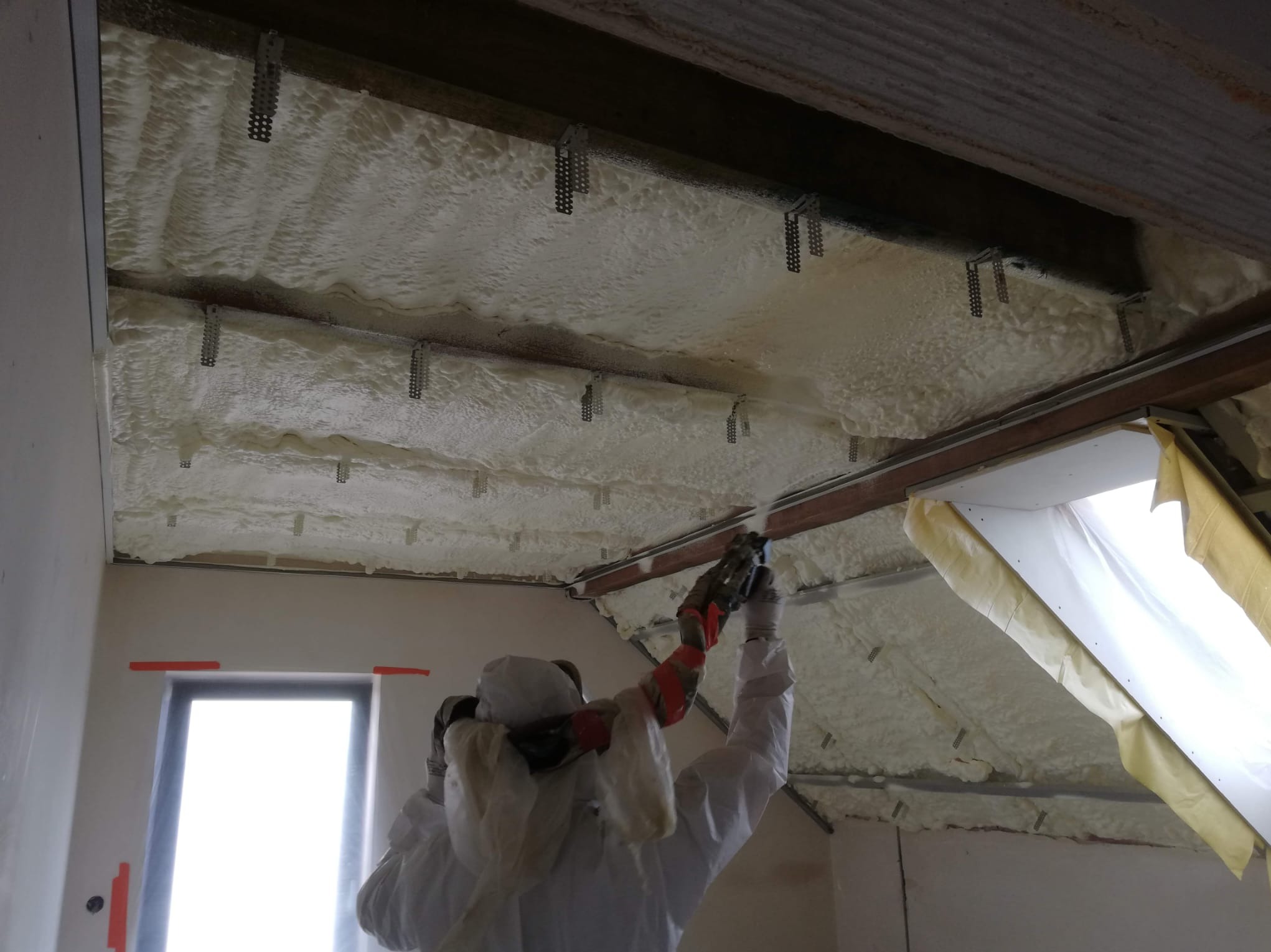 Prace związane z ocieplaniem poddasza pianką PUR w Polkowicach. Widoczny pracownik w kombinezonie ochronnym aplikujący pianę na konstrukcję dachu.