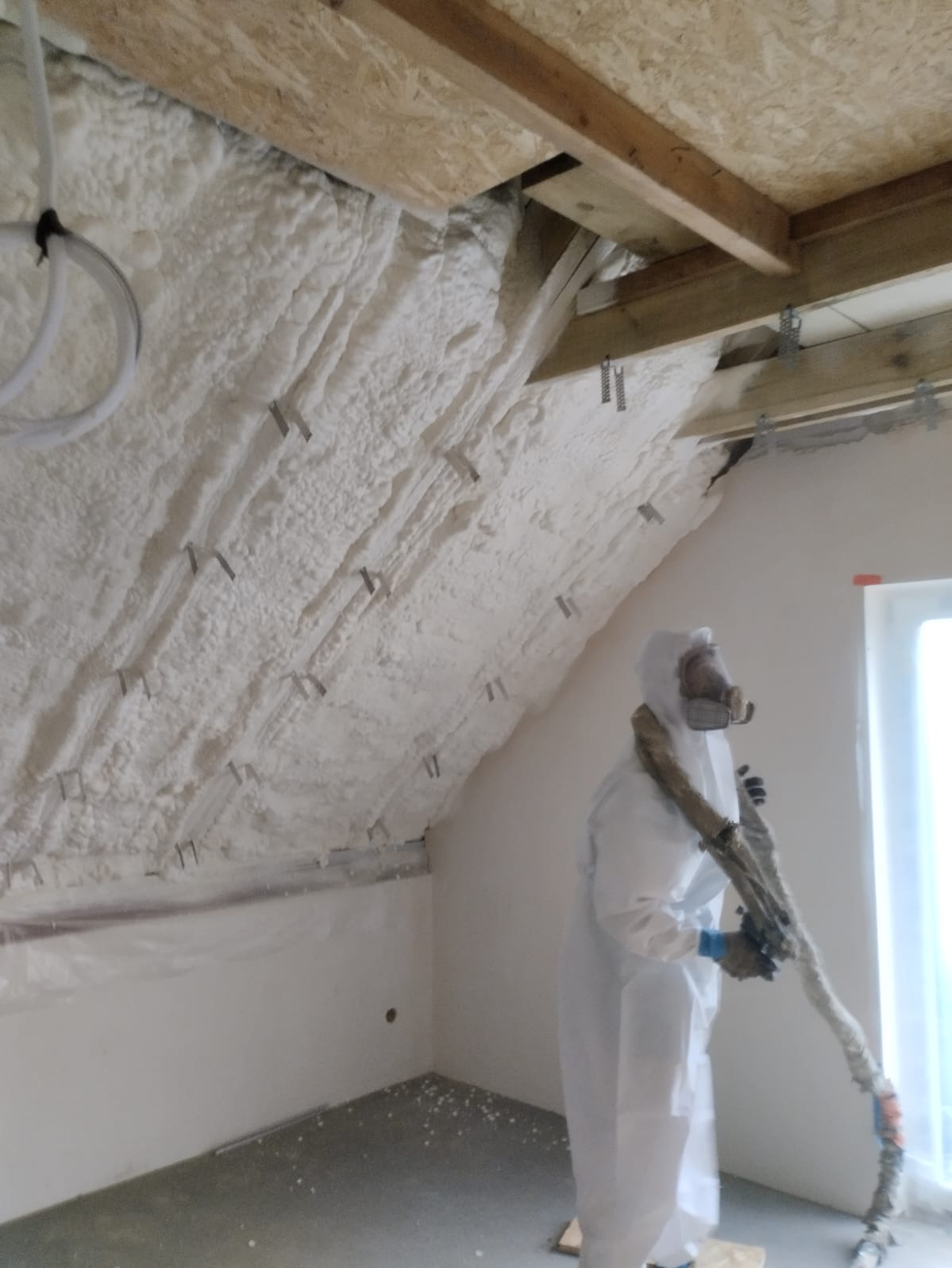 Pracownik w kombinezonie ochronnym aplikuje białą pianę izolacyjną na skośny strop poddasza, widoczne metalowe uchwyty mocujące, surowe drewniane belki i fragment okna.