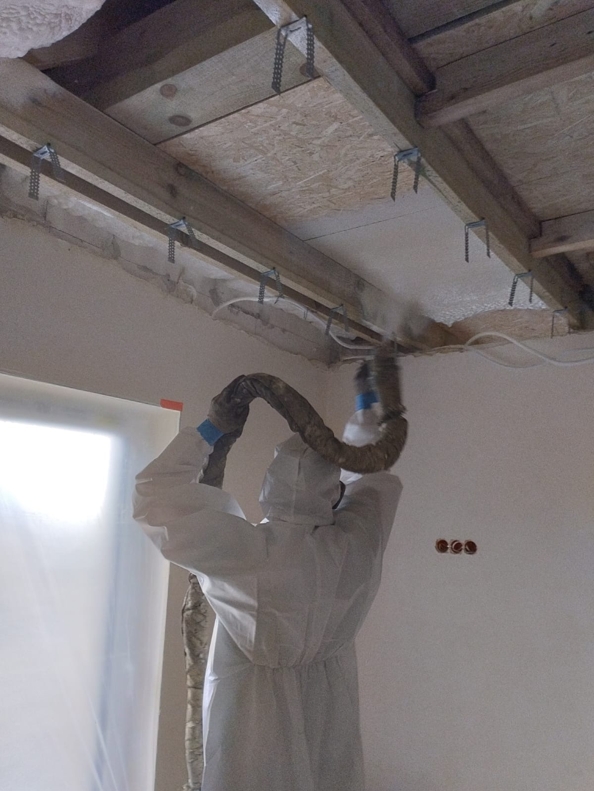 Pracownik w kombinezonie ochronnym aplikuje izolację natryskową na poddasze, widoczne drewniane elementy konstrukcyjne dachu i mocowania.