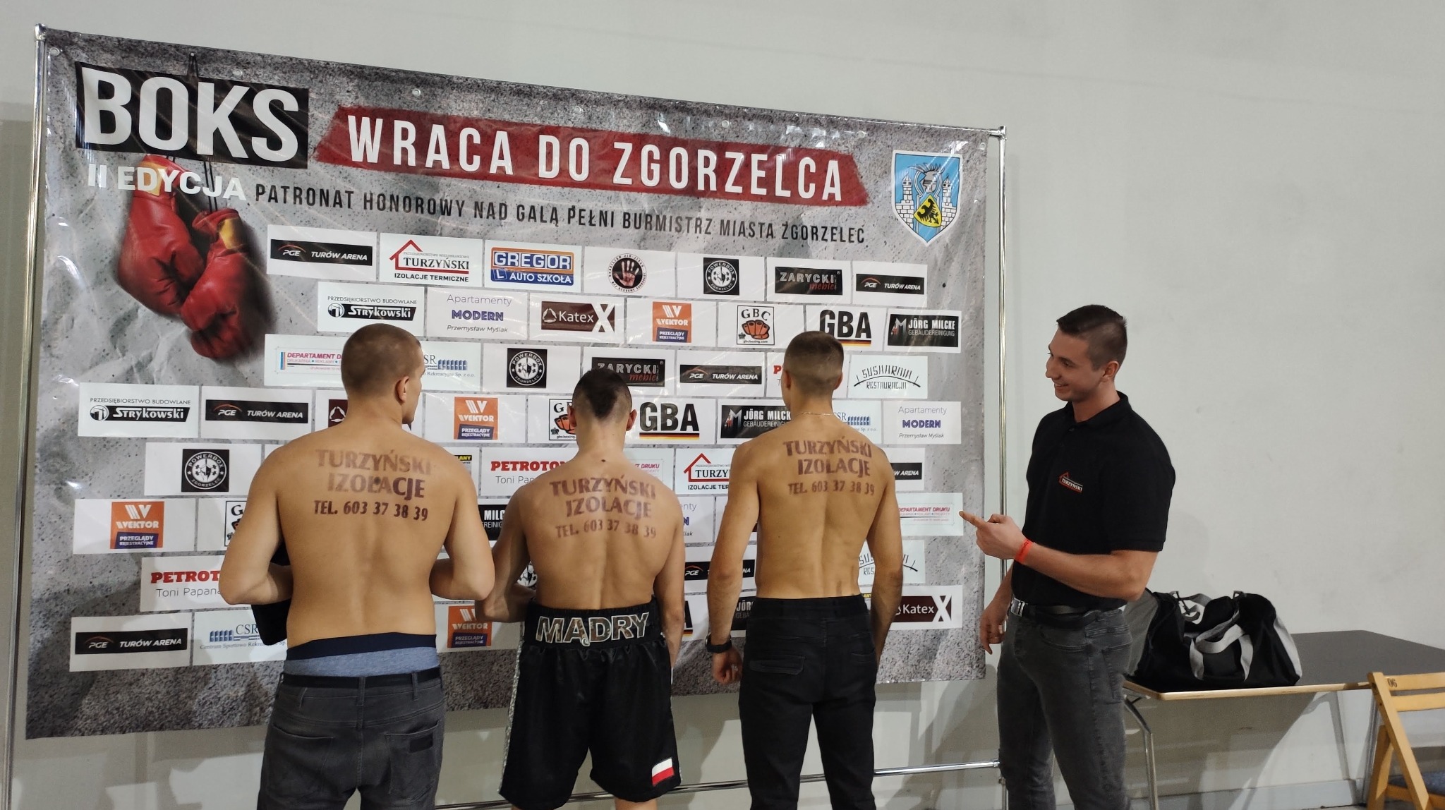 Trzej mężczyźni z nagimi plecami, na których wypisano 'Turzyński Izolacje TEL. 603 37 38 39', stoją przed banerem reklamowym z logo firmy i hasłem 'Boks wraca do Zgorzelca', obok nich stoi...