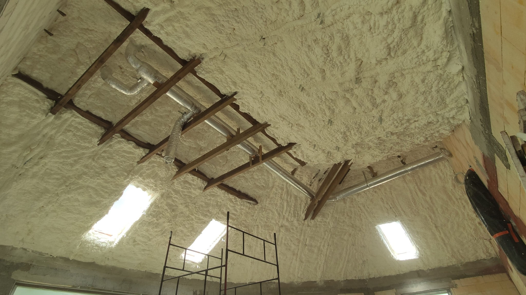 Poddasze z widoczną konstrukcją drewnianą, pokryte grubą warstwą jasnej piany izolacyjnej, z fragmentami instalacji wentylacyjnej i oknami dachowymi, widoczne rusztowanie.