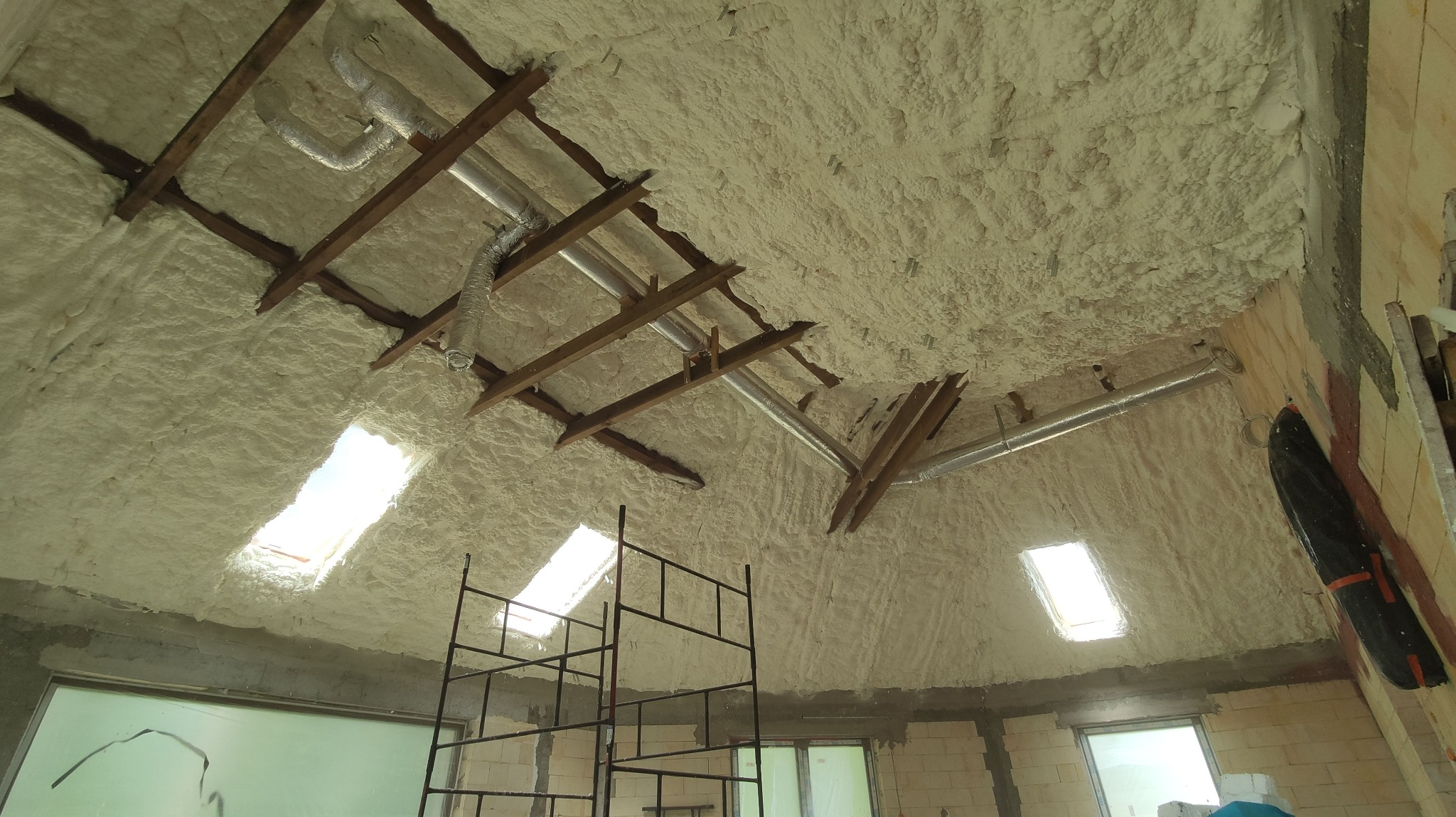 Poddasze z drewnianą konstrukcją i instalacją wentylacyjną pokryte warstwą izolacyjną z widocznymi oknami dachowymi oraz fragmentem rusztowania.