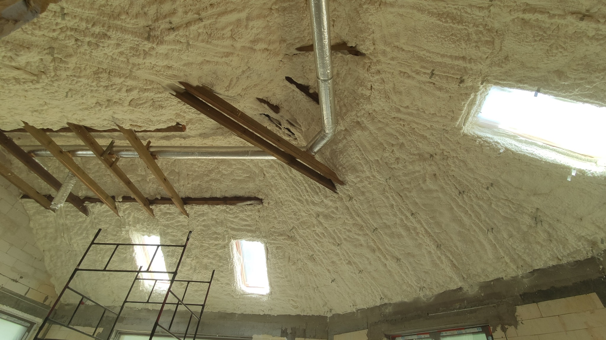 Poddasze z widoczną konstrukcją drewnianą i metalowymi rurami wentylacyjnymi, pokryte grubą warstwą jasnej piany izolacyjnej, widoczne okna dachowe i rusztowanie