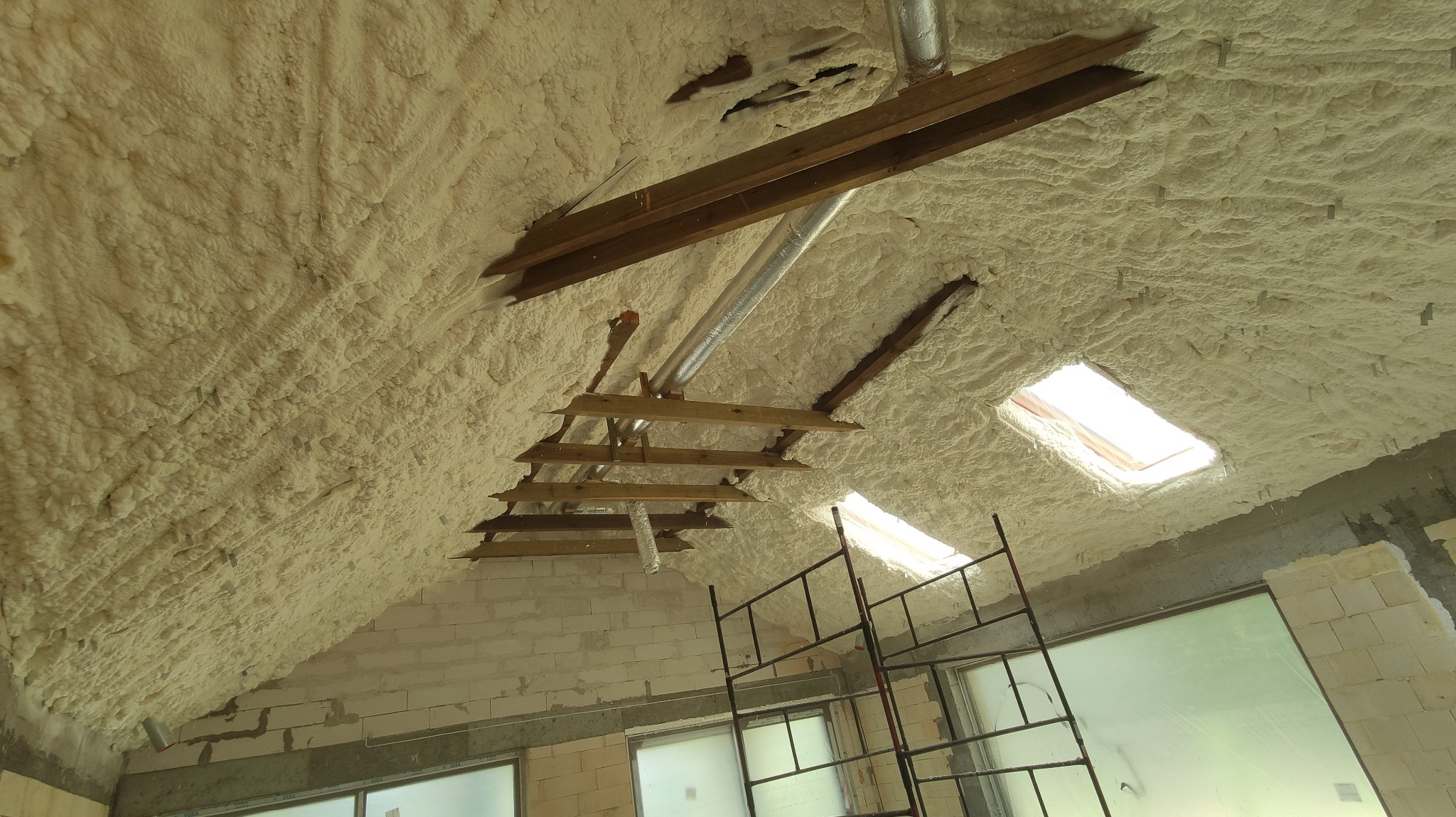 Poddasze z widoczną izolacją natryskową, drewnianą konstrukcją, metalowymi rurami wentylacyjnymi i oknami dachowymi, widok z dołu, na ścianach widoczne białe bloczki.