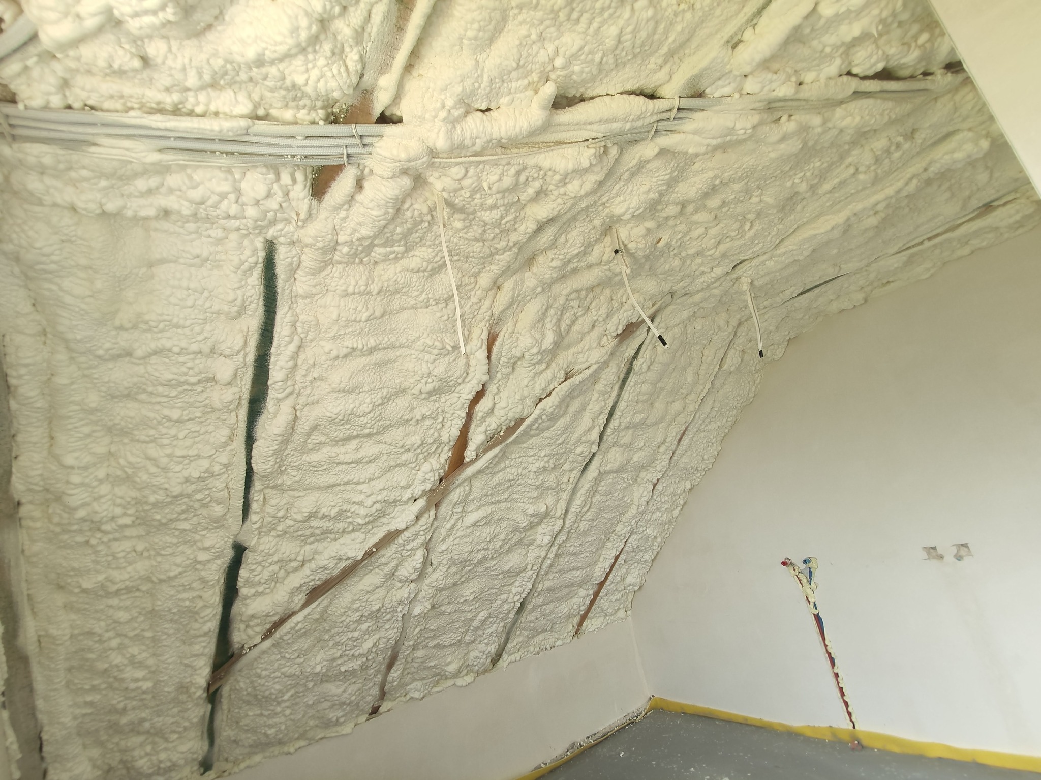 Poddasze z widoczną, nierównomierną warstwą białej piany izolacyjnej, częściowo zakrywającej drewniane elementy konstrukcyjne i kable, z fragmentem ściany z wystającymi przewodami elektrycznymi w tle.