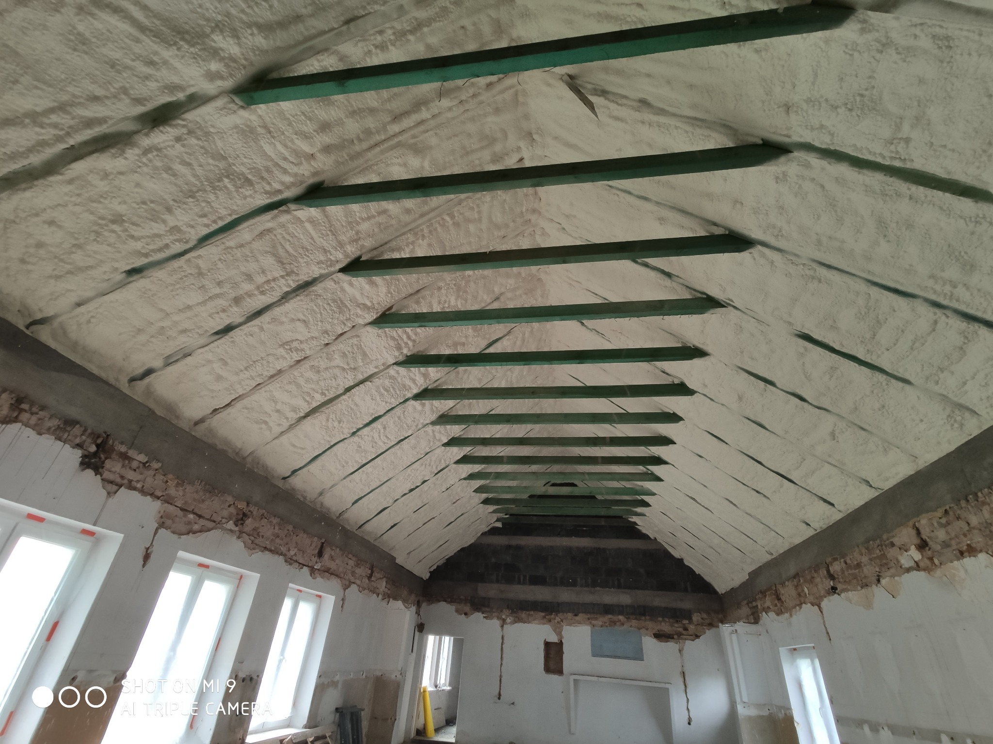 Ocieplone pianką PUR poddasze z widocznymi zielonymi belkami konstrukcyjnymi, surowe ściany w trakcie remontu z oknami w tle.