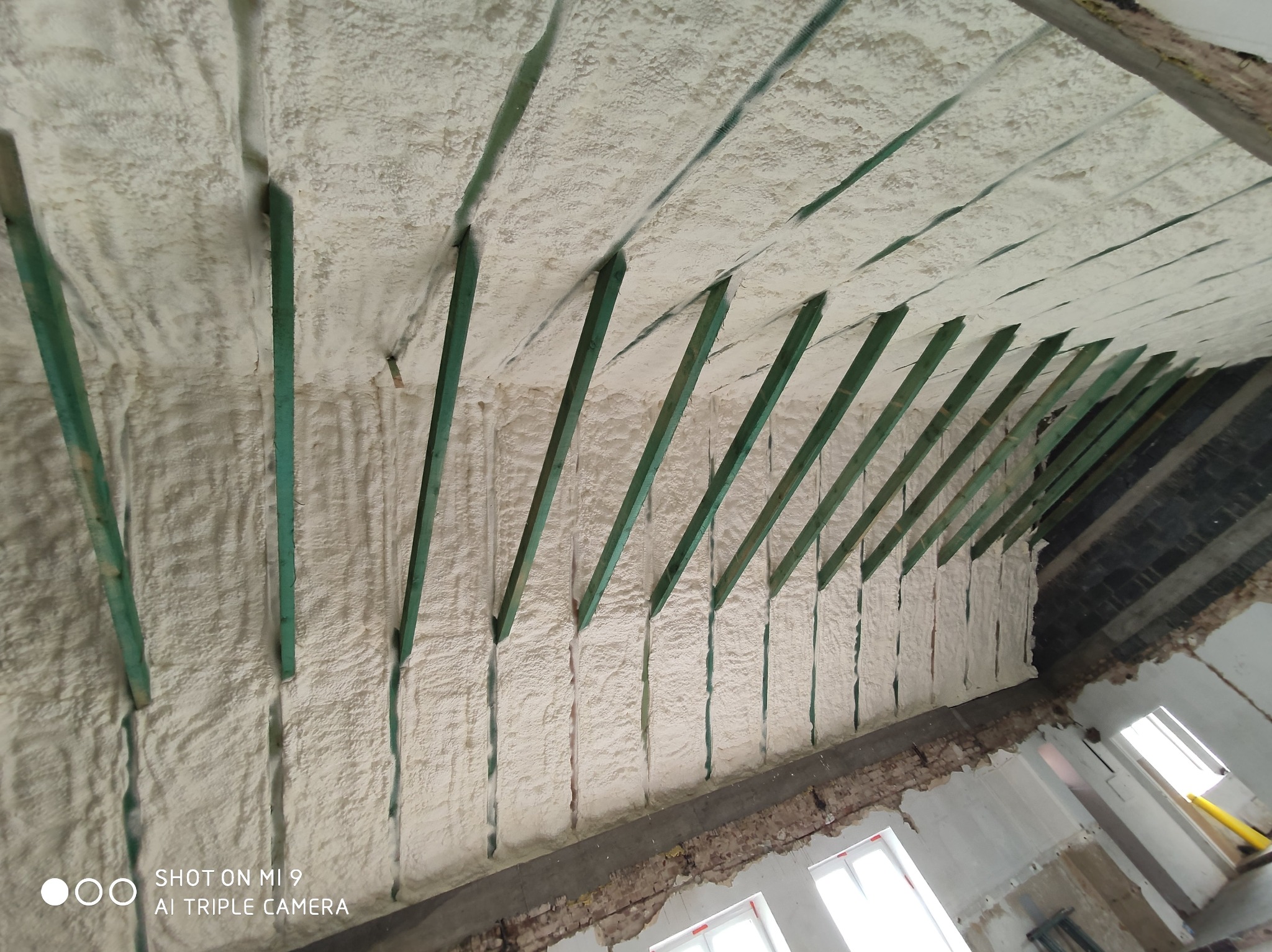 Poddasze z drewnianą konstrukcją krokwiową wypełnione białą, spienioną izolacją, widoczne fragmenty muru i okien w stanie surowym.