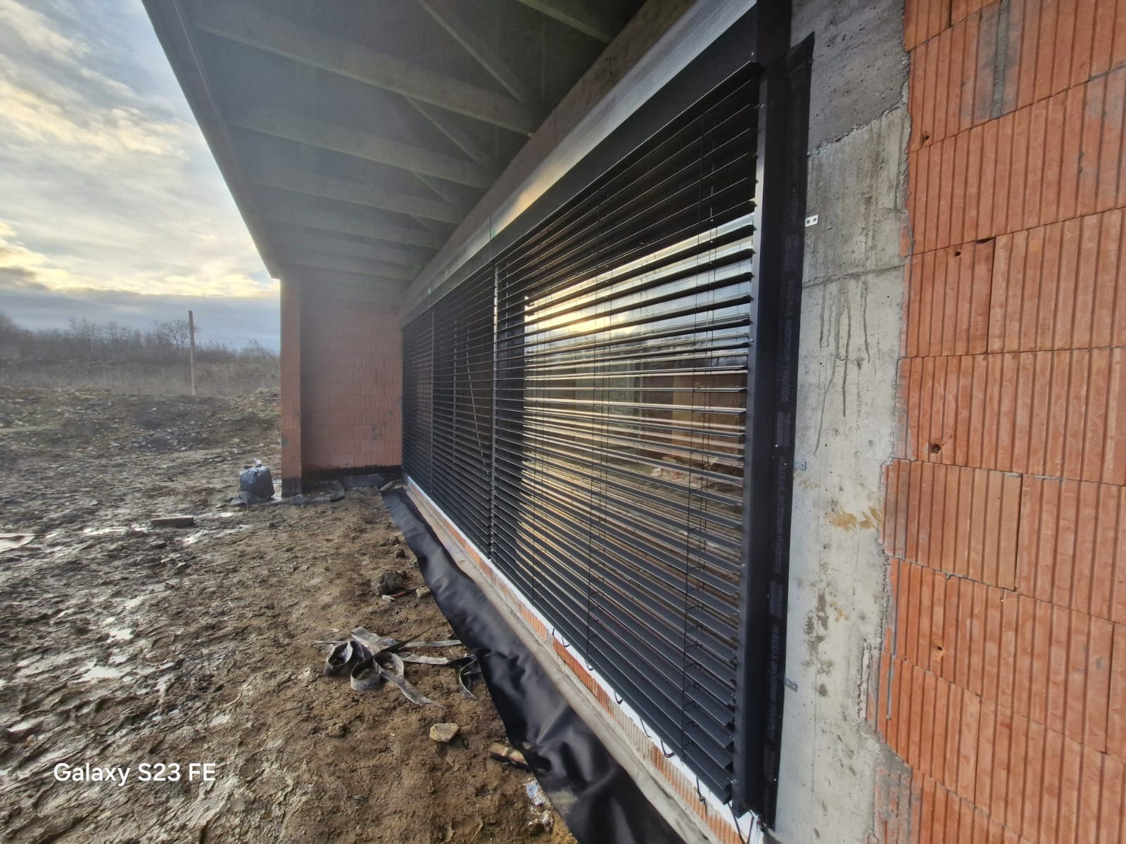 Ciemne żaluzje fasadowe zamontowane w surowym budynku z czerwonej cegły, widoczny fragment dachu i niezagospodarowany teren.