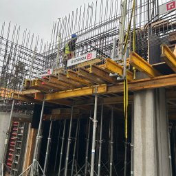 kkBudowlana - Konstrukcja zbrojeniowa z deskowaniem i podporami na budowie, pracownik w kasku i kamizelce odblaskowej na górze, widoczne logo firmy na deskowaniu.