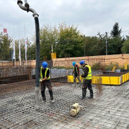 kkBudowlana - Betonowanie płyty fundamentowej zbrojonej siatką stalową; pracownicy w kaskach i kamizelkach odblaskowych w trakcie wylewania betonu z pompy; w tle kominy fabryczne.