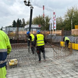 kkBudowlana - Prace betoniarskie: robotnicy w kaskach i kamizelkach wylewają beton z pompy na przygotowane zbrojenie, widoczny element szalunku i tło z budynkami oraz kominem.