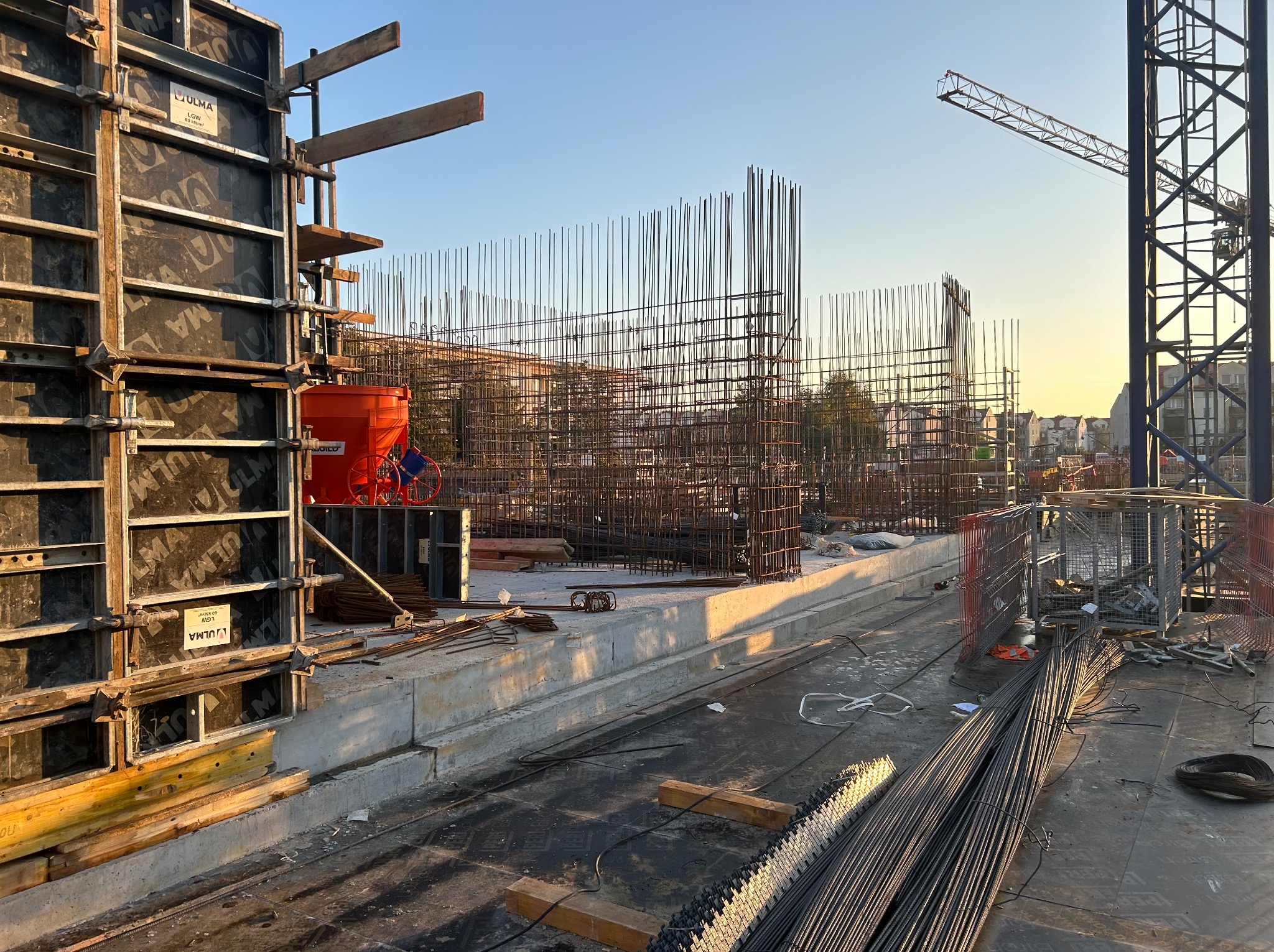 Konstrukcja zbrojeniowa szkieletu budynku w trakcie budowy, widoczne szalunki ULMA, stalowe pręty zbrojeniowe, pomarańczowy zasobnik na beton oraz żuraw budowlany na tle błękitnego nieba...