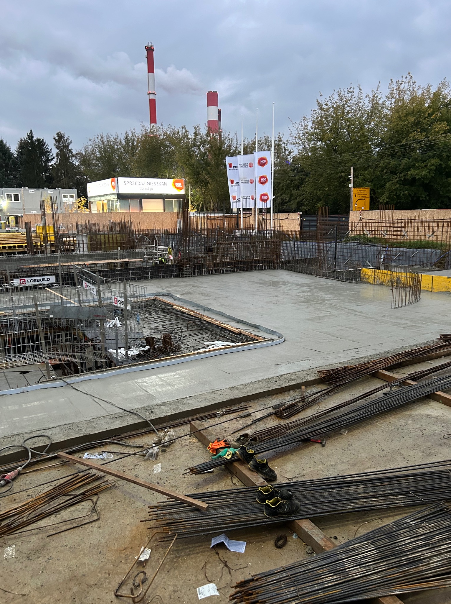 Wylany fundament żelbetowy z widocznym zbrojeniem, w tle kominy i budynek biura sprzedaży mieszkań.
