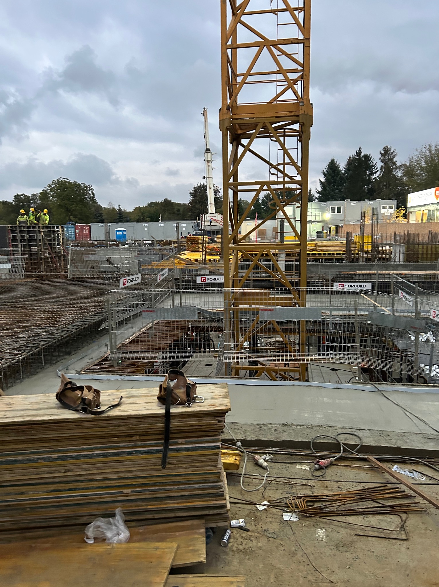 Plac budowy z żółtym dźwigiem, widoczne szalunki, zbrojenia, beton, paleta desek z narzędziami, pracownicy w żółtych kamizelkach na podwyższeniu.