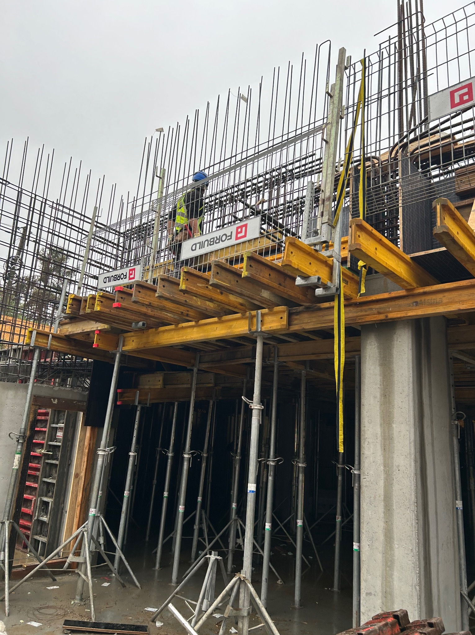 Konstrukcja zbrojeniowa z deskowaniem i podporami na budowie, pracownik w kasku i kamizelce odblaskowej na górze, widoczne logo firmy na deskowaniu.