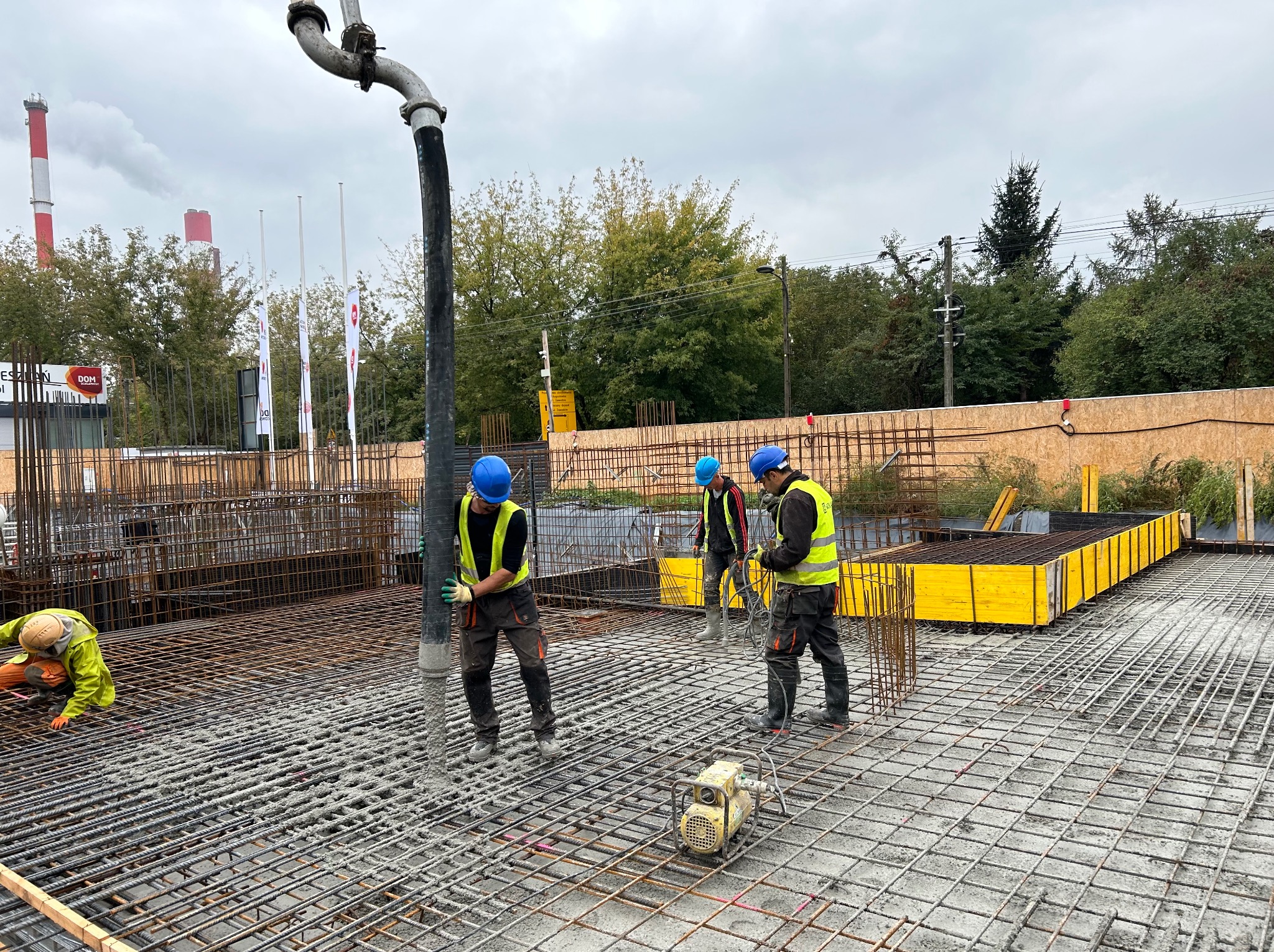 Betonowanie płyty fundamentowej zbrojonej siatką stalową; pracownicy w kaskach i kamizelkach odblaskowych w trakcie wylewania betonu z pompy; w tle kominy fabryczne.