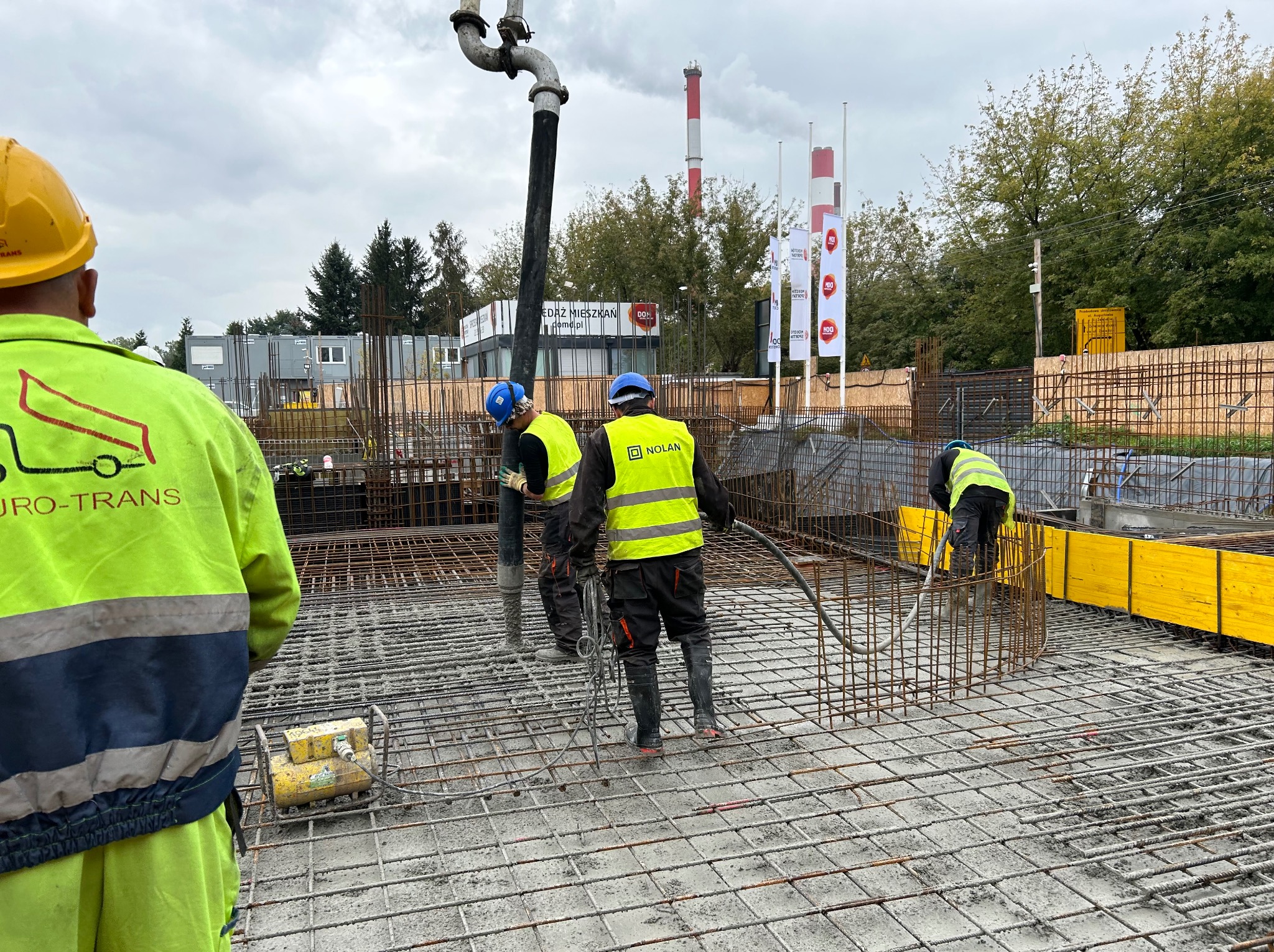 Prace betoniarskie: robotnicy w kaskach i kamizelkach wylewają beton z pompy na przygotowane zbrojenie, widoczny element szalunku i tło z budynkami oraz kominem.