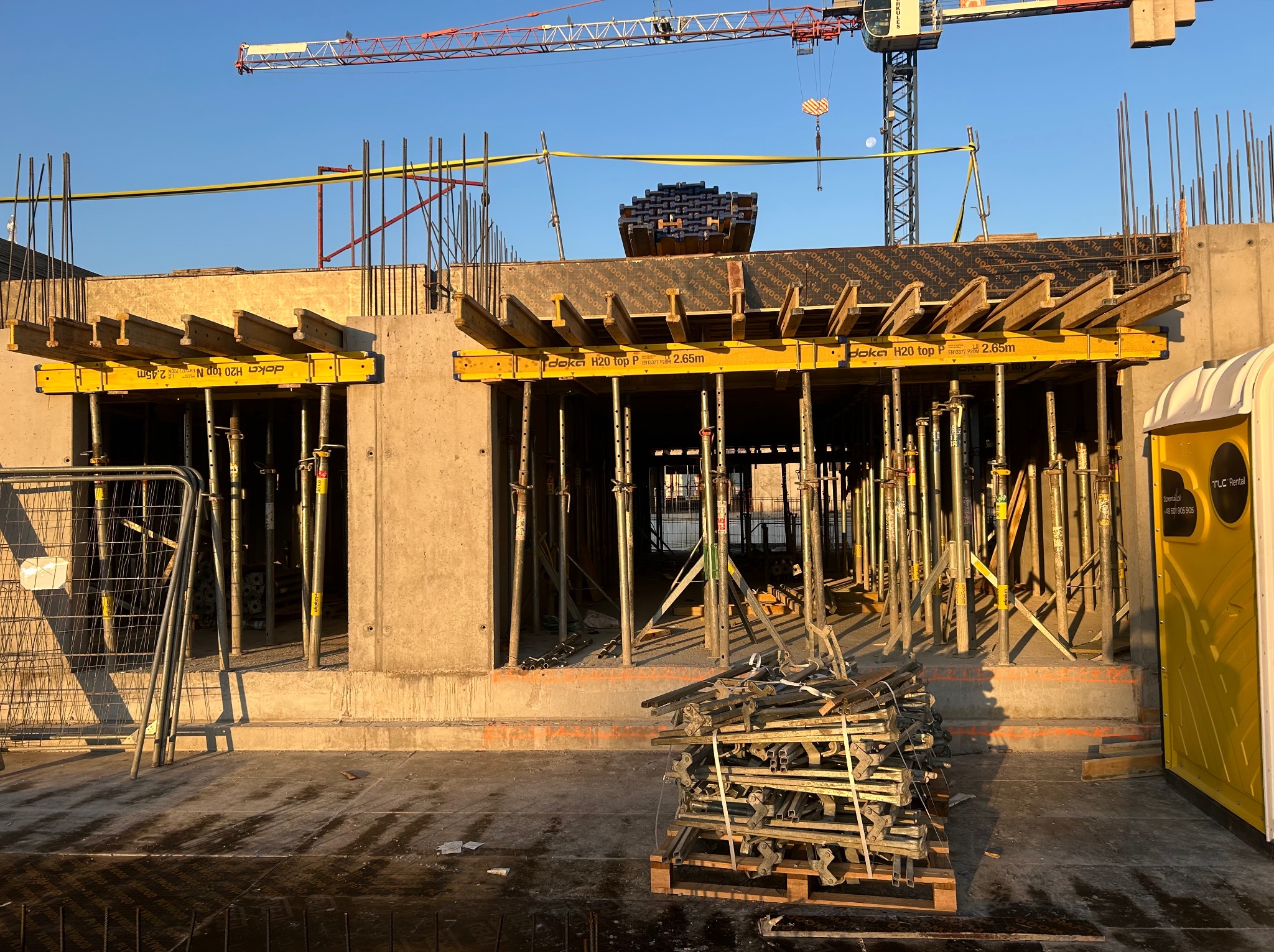 Wznoszenie konstrukcji żelbetowej: szalunki stropowe Doka H20 top, stalowe podpory i widoczny dźwig budowlany na tle błękitnego nieba.