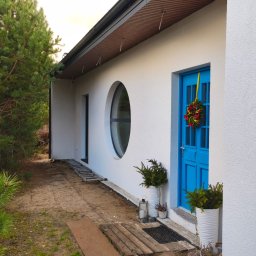 HH - Happy House Sebastian Chrzanowski - Biały budynek z okrągłym oknem i niebieskimi drzwiami ozdobionymi wieńcem, obok rosną drzewa iglaste, na ziemi leży drabina.