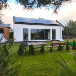 HH - Happy House Sebastian Chrzanowski - Nowoczesny, parterowy dom z białymi ścianami i panelami słonecznymi na dachu, widok z ogrodu z zielonym trawnikiem, drewnianym stołem i ławką oraz młodymi drzewkami iglastymi.