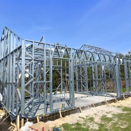 HH - Happy House Sebastian Chrzanowski - Konstrukcja domu ze stalowym szkieletem na betonowej płycie fundamentowej, widok na drzewa w tle, pracownik na górze konstrukcji.