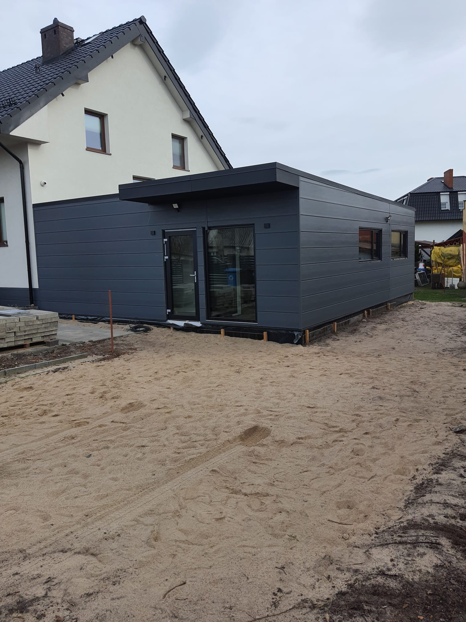 Nowoczesna, grafitowa dobudówka w Luzinie z płaskim dachem i panelami elewacyjnymi, widoczna na tle piaszczystego terenu i istniejącego domu.